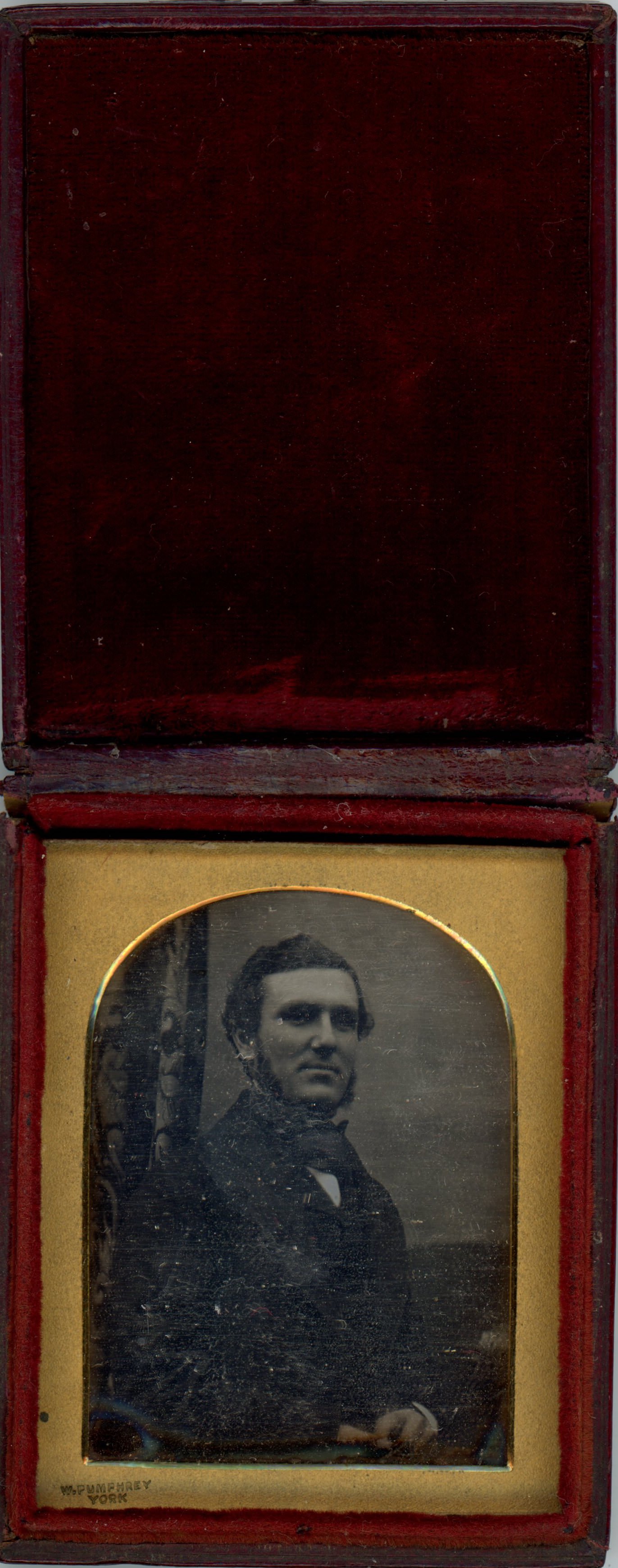 Daguerreotype. A pale-skinned man with mutton chops and dark hair looks at us with deep-set eyes and a hint of a smile. The gold mat has an arched top. The red case is flipped up and the underside of the lid is covered in dark red cloth.