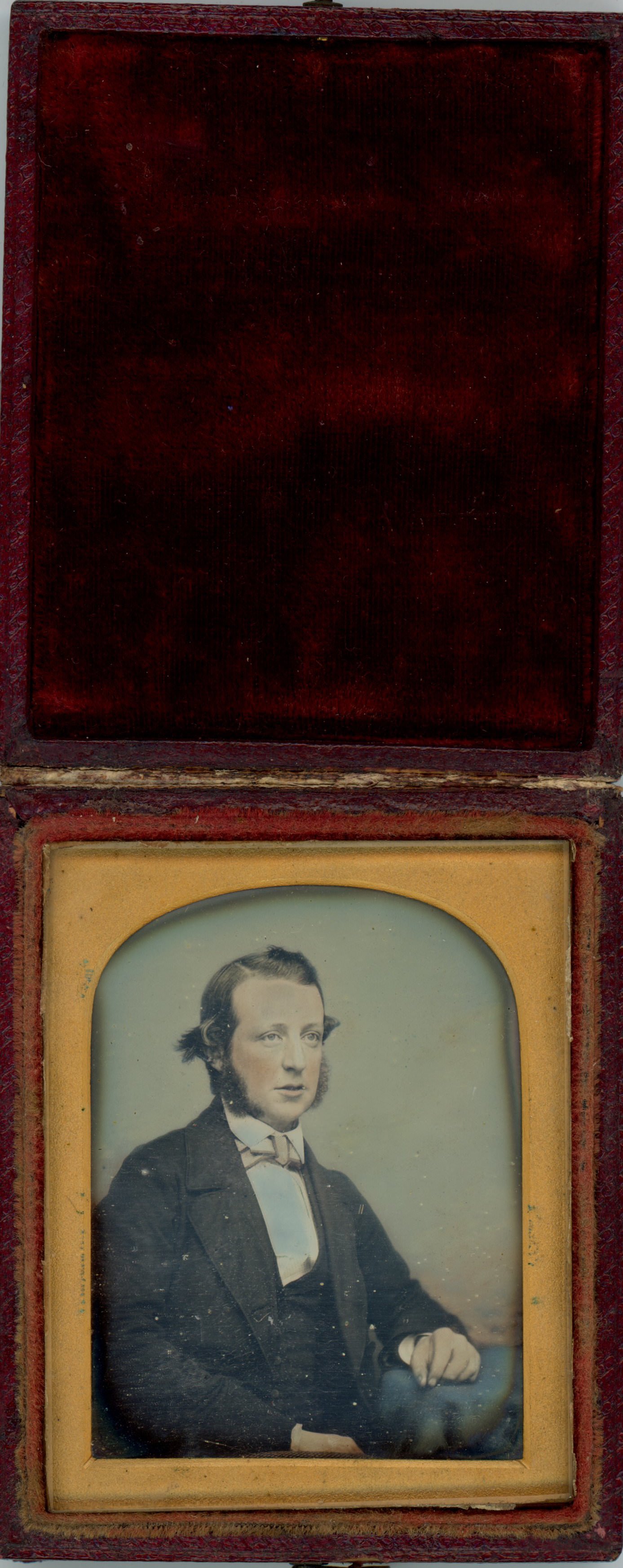 Daguerreotype. A pale-skinned man with mutton chops looks off to our right. His hair flares out over his ears, and his lips are parted. The photograph has been hand-coloured with peach tone for the skin, pink for the cheeks, and blue for the background and props.