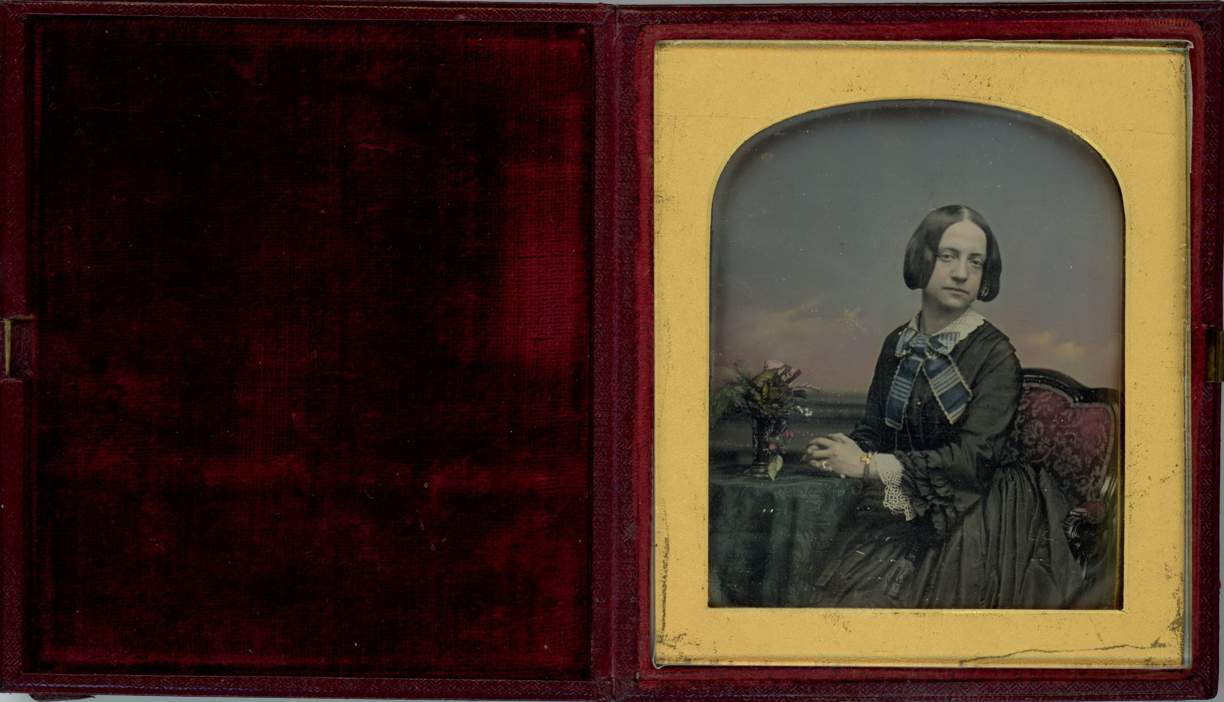 Daguerreotype. A pale-skinned woman wearing a black dress looks at us. Her hands are clasped and rest on a table holding a spray of flowers in a vase. Hand-colouring has been added to the props, background, clothing, and her skin.