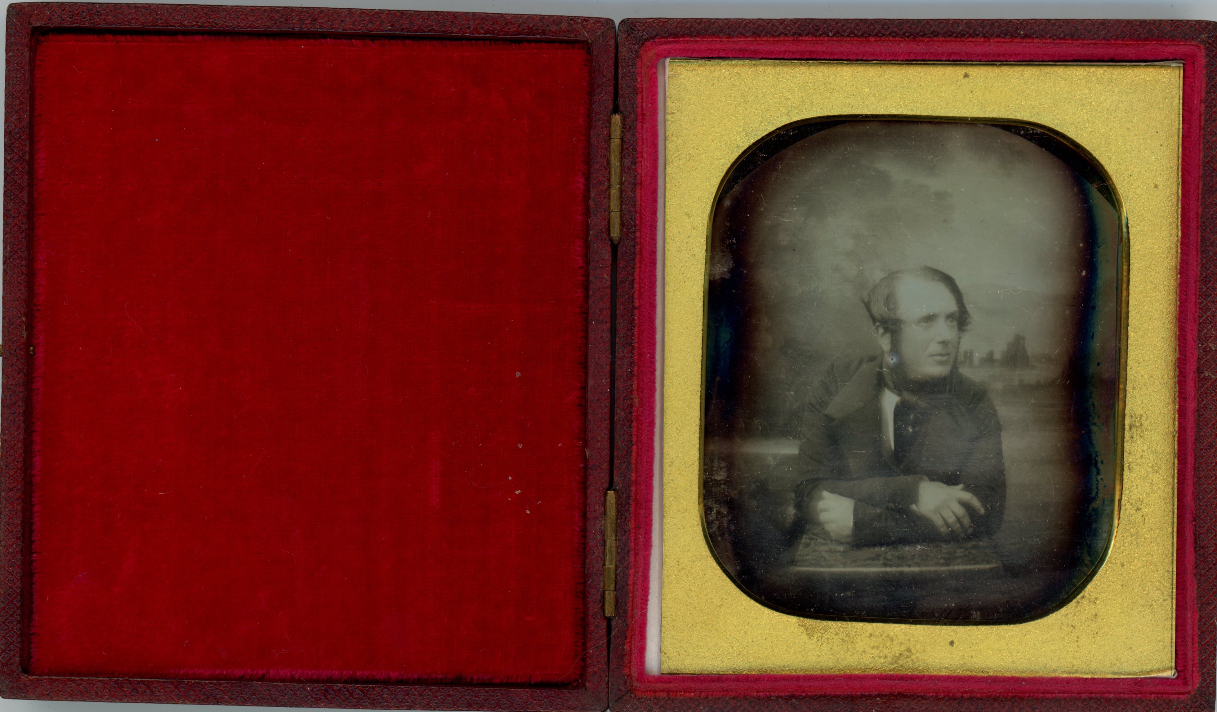 Daguerreotype. A pale-skinned man with mutton chops leans forwards onto his crossed arms as he looks off to our right. His dark hair is swept forwards over his ears, and his lips are parted. The background shows a distant landscape.