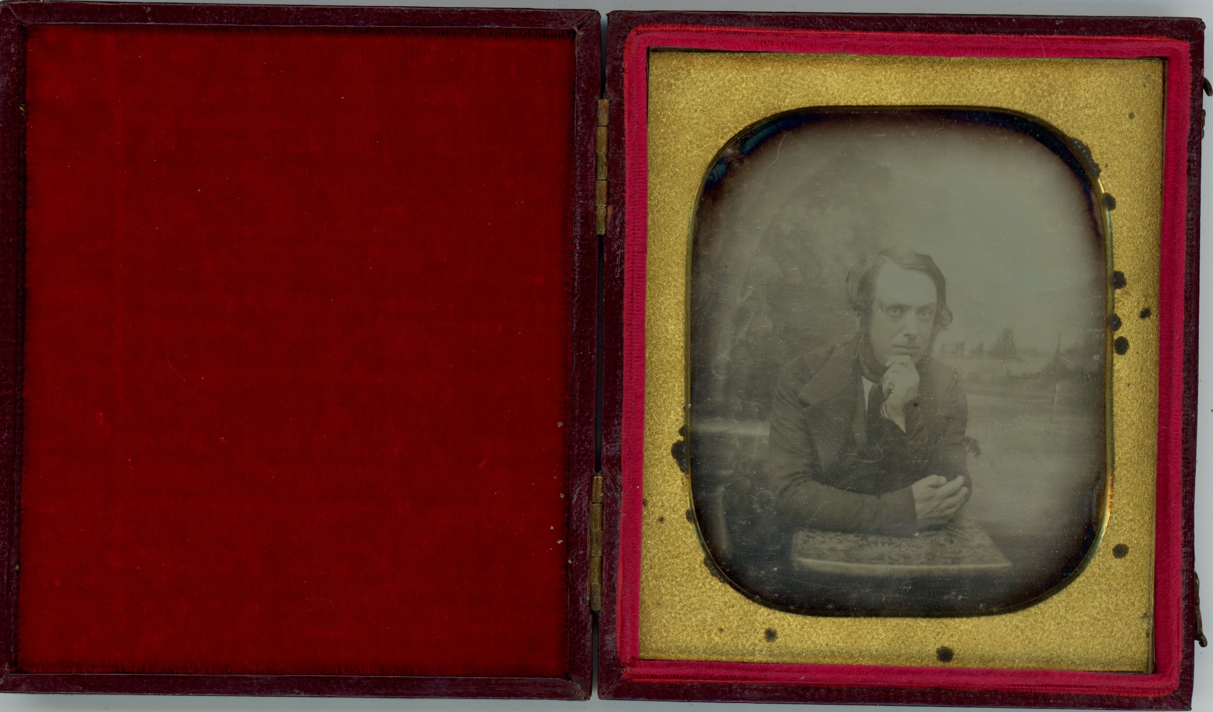 Daguerreotype. A pale-skinned man with mutton chops leans forwards onto his elbows, his chin resting in one hand. He looks at us from in front of a distant landscape background. The gold mat is rounded at the corners.