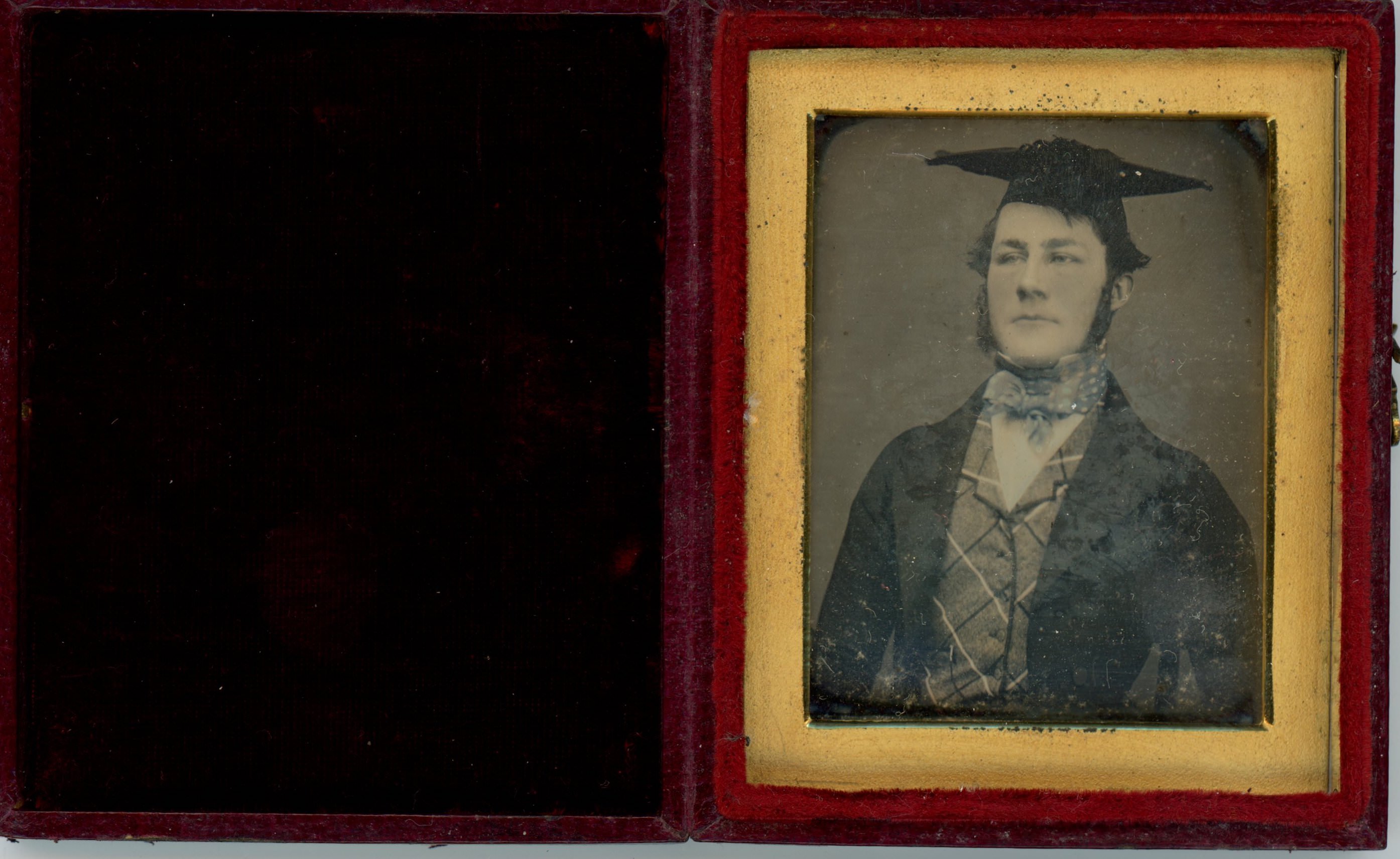 Daguerreotype in an open case. A pale-skinned young man wearing a mortar board cap looks stiffly off to our left. He wears a vest, jacket, and a cravat coloured light blue. The photograph is in a gold frame in a red case, which opens like a book. The cover is lined with black fabric.