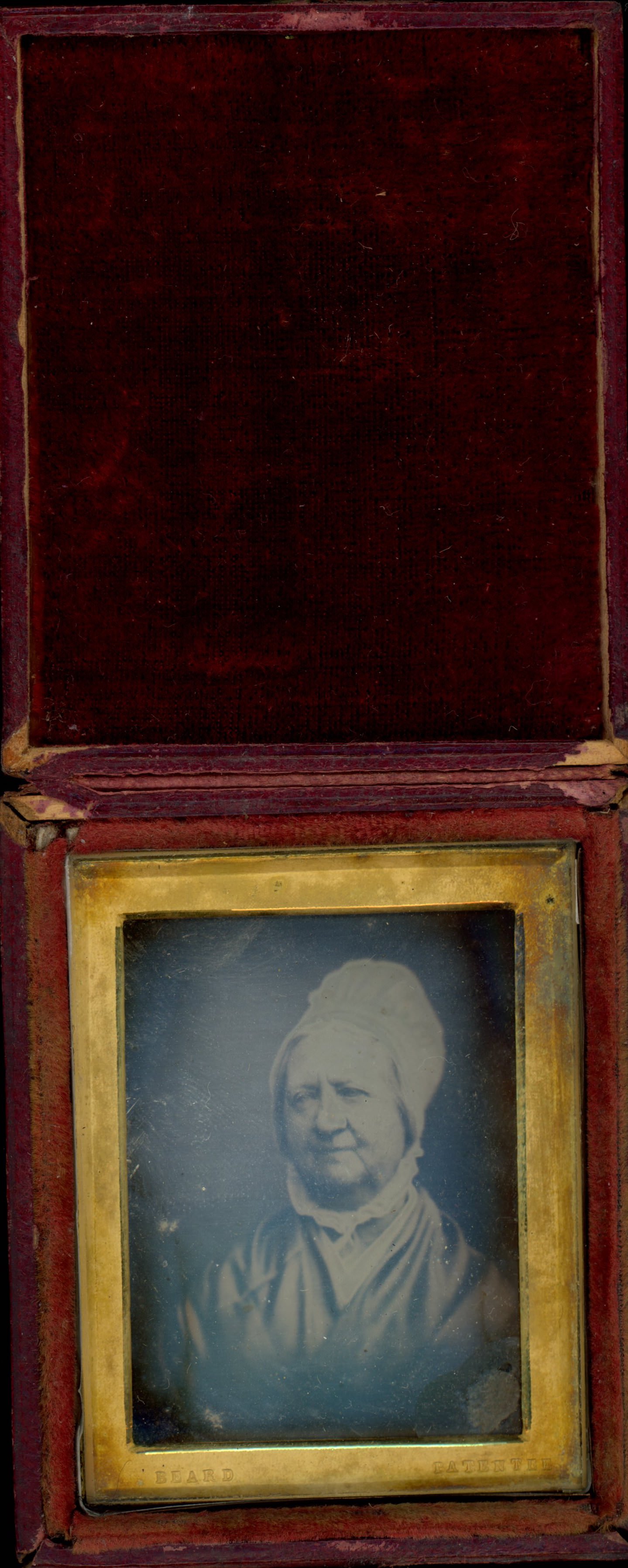 Daguerreotype in an open case. A pale-skinned woman with rounded cheeks and sagging jowls looks off to our left in a rectangular gold mat set in a red case. The words “Beard” and “Patentee” are stamped into the bottom corners of the gold mat.