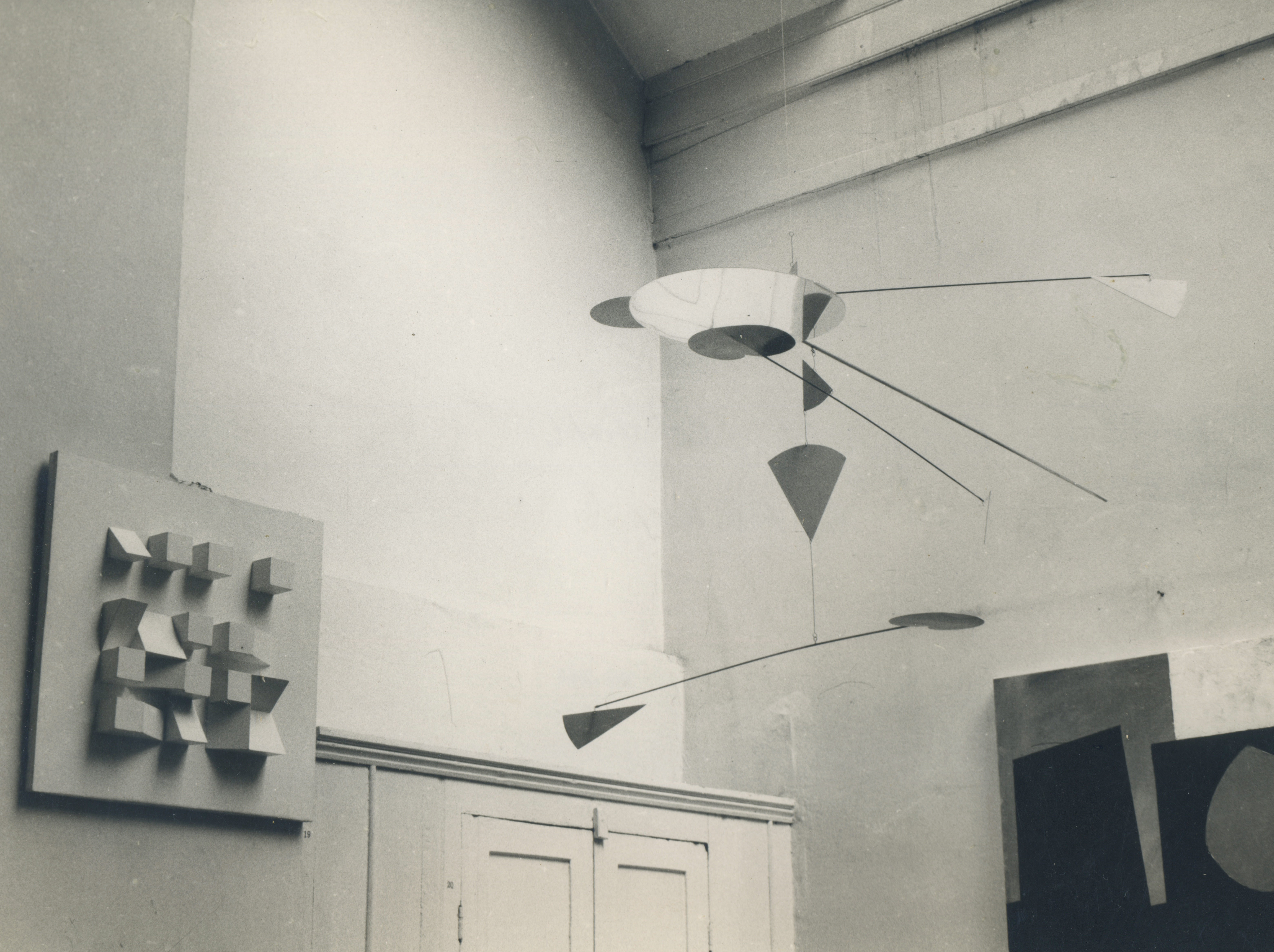 Installation photograph. In this black and white photograph, sculptural works are displayed a high-ceilinged room. To our left is a flat white square from which sixteen cubes, triangles, and other polygonal blocks project in a grid. A mobile with fine arms that suspend flat geometric shapes hangs from the ceiling. The edge of another abstract work is visible in the lower right corner of this view.