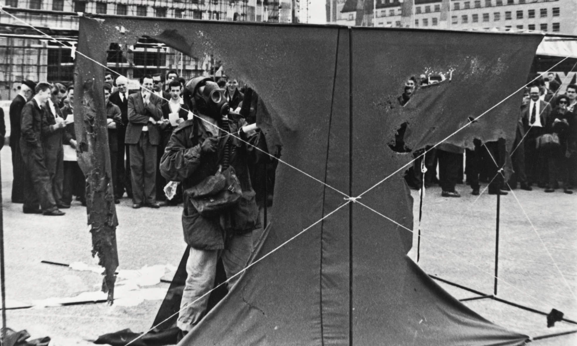 Performance art. A piece of dark cloth the size of a single car garage is stretched across two rails, one above and one below, in this black and white photograph. From the far side of the cloth, we face the artist, who wears a gas mask and military jacket as he sprays acid onto the cloth. Large swaths have been chewed away, leaving an inwards-curving, wide, T shape of fabric behind.
