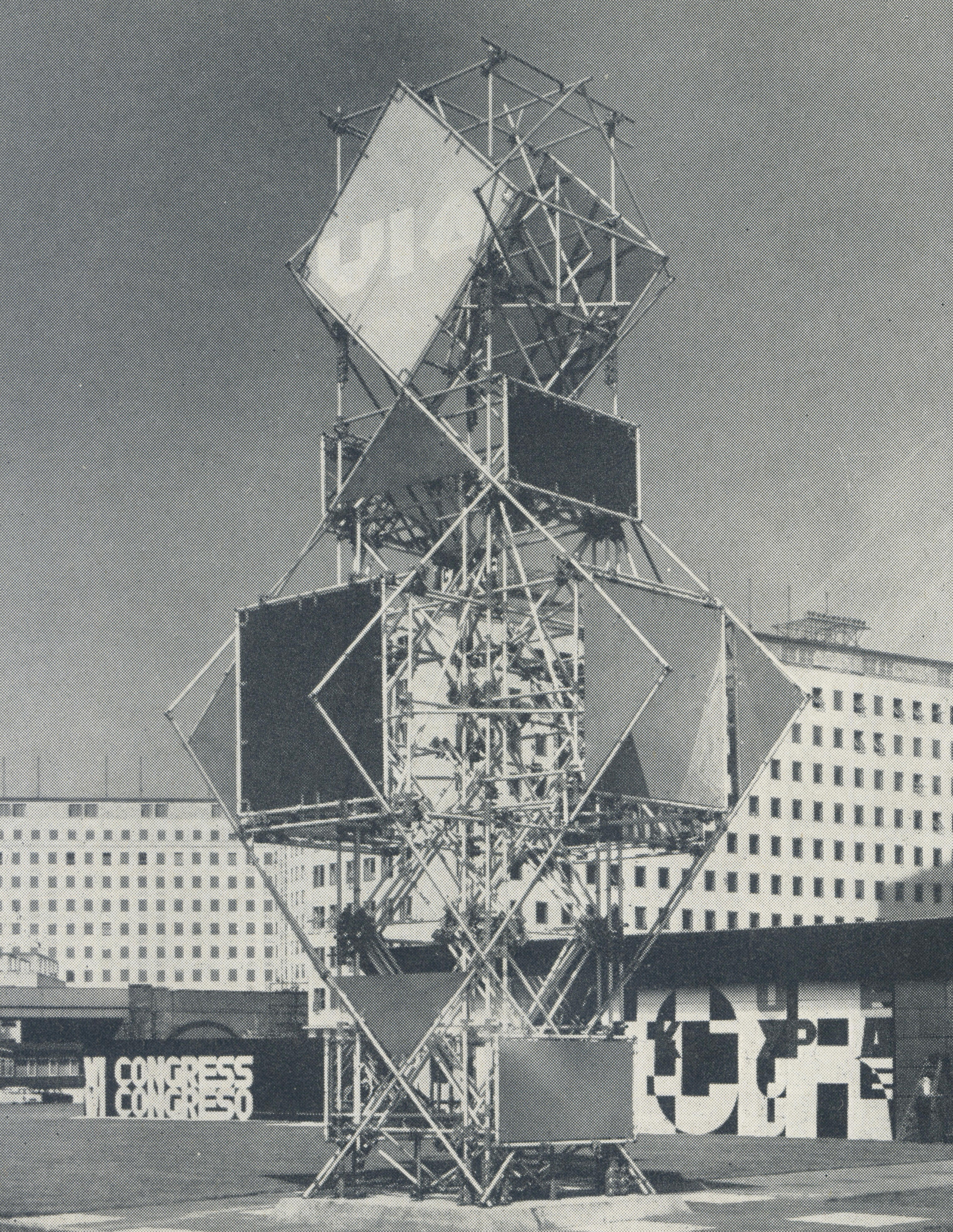 Black and white photograph. A sculptural tower is made of scaffolding built into a large diamond shape under a smaller one above. Some square and triangular panels are set against the scaffolding. It appears to be several storeys tall against the buildings in the background.