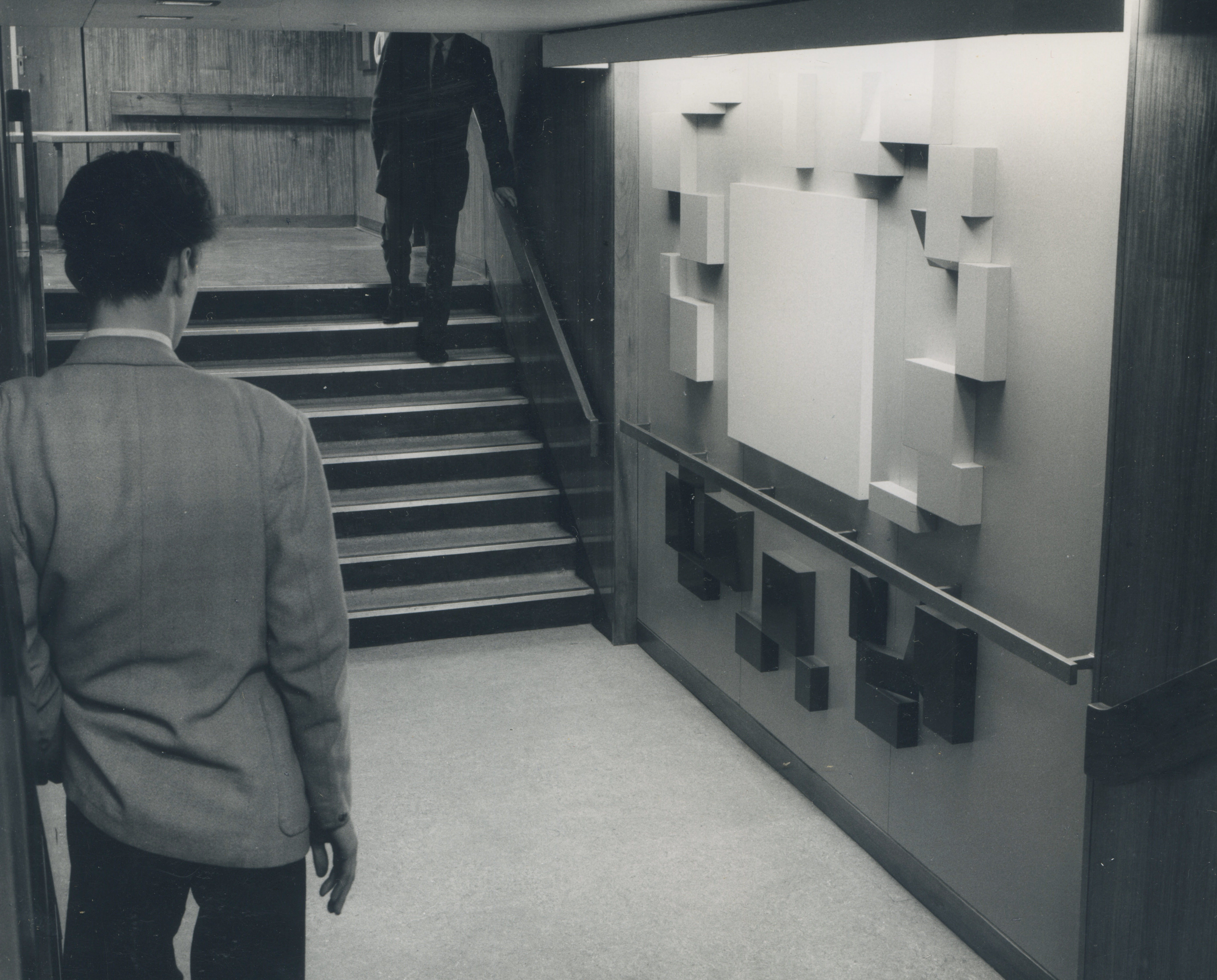 Black and white photograph. Dark and light squares, rectangles, and panels are installed on a wall above and below a railing. The panels above the railing are lighter in tone than those below. A person stands near us, facing away and looking towards the installation. A half-flight of stairs is just beyond the railing.