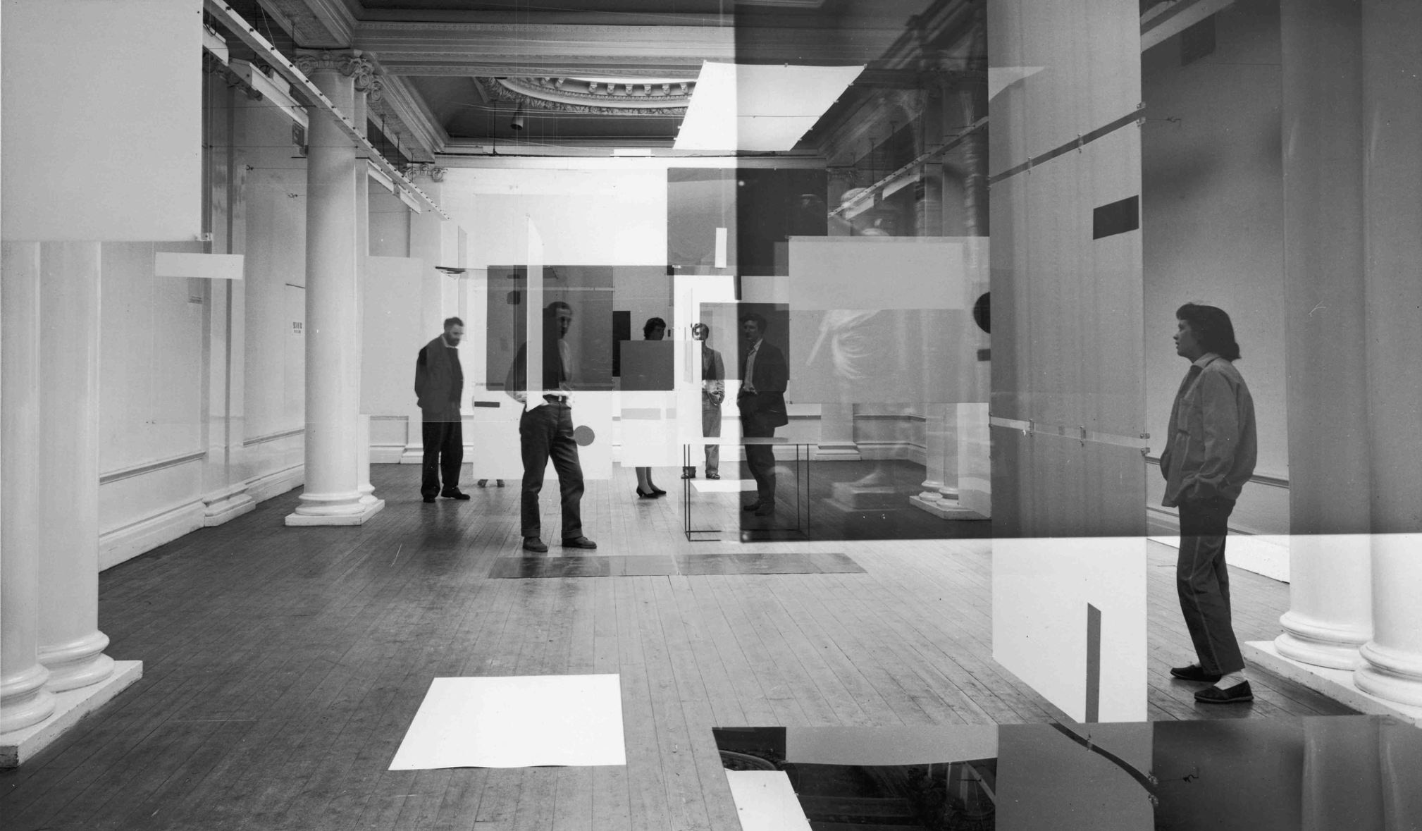 Installation photograph. Light and dark translucent panels hang in a gallery space between us and people standing in the room. The screens hanging from the ceiling and reflect or obscure people, creating a confused, surreal space.