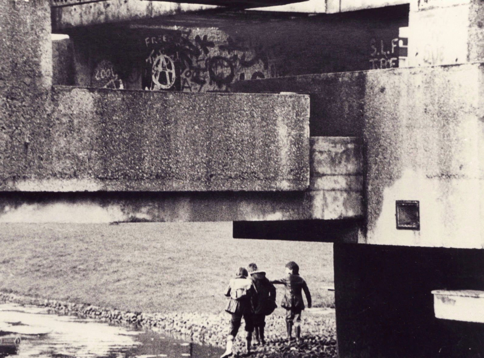 Architecture. In this black and white photograph, concrete planes and ledges make up a bewildering space. Three children stand by water at the concrete edge of an aqueduct.