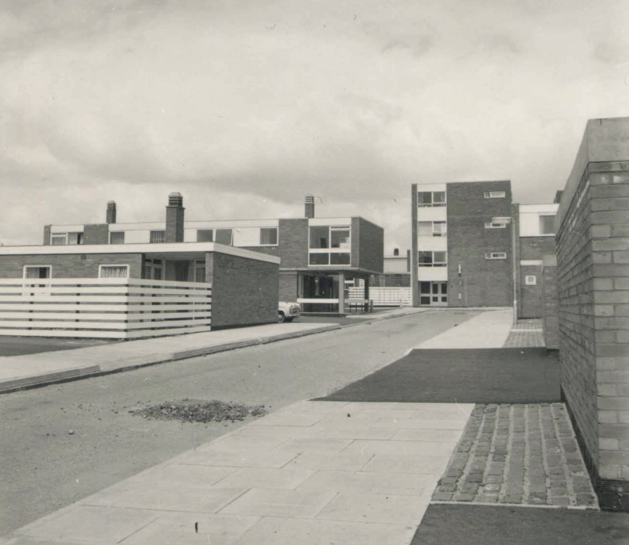 Architecture. Black and white photograph of blocky residential buildings and horizontal fencing. The bildings are between one and four stories high. A street that recedes across the image and its sidewalks are empty.