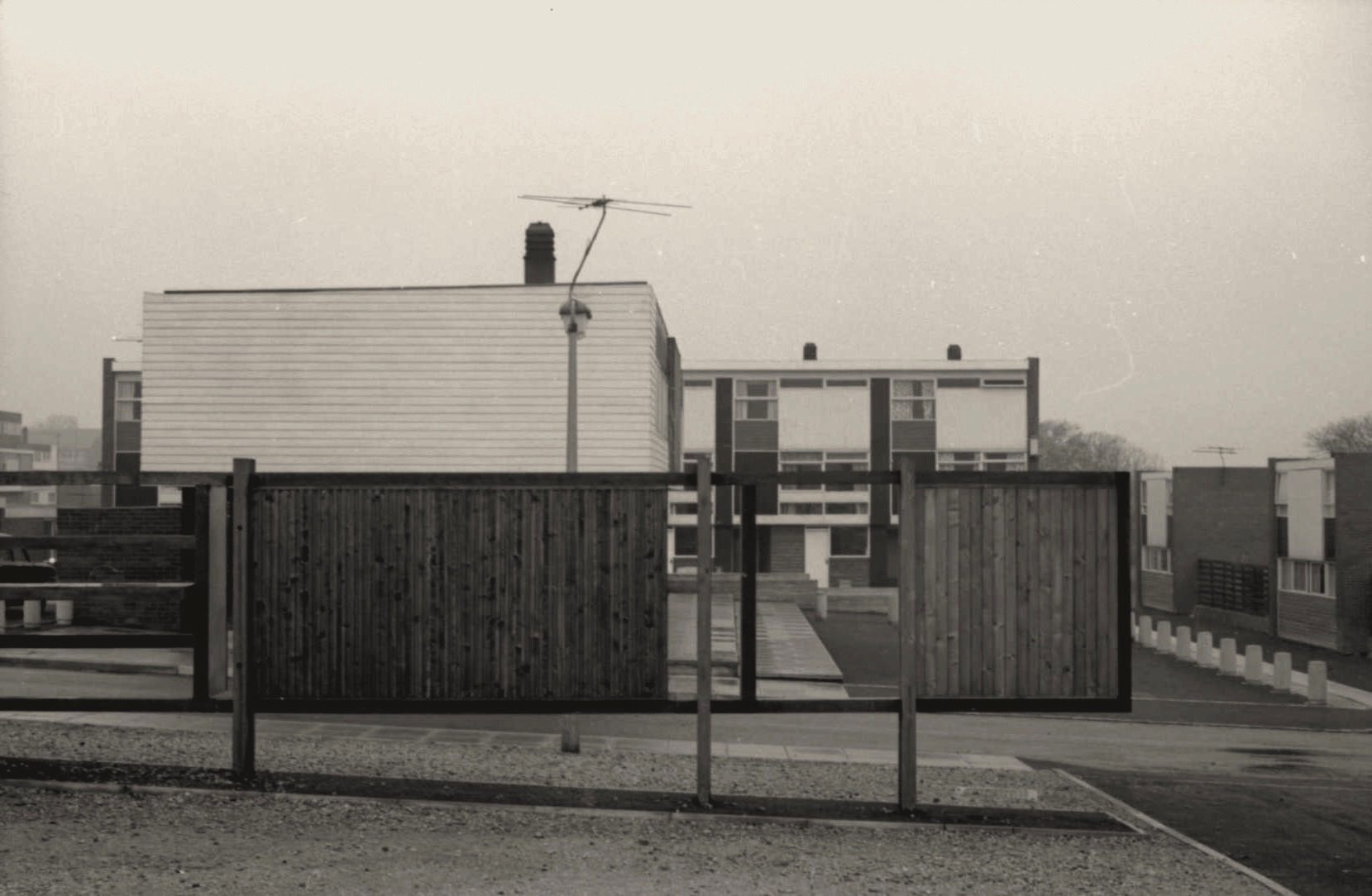 Architecture. Panels of vertically oriented fencing and horizontal boards are against blocky buildings with rectangular windows in this black and white photograph.