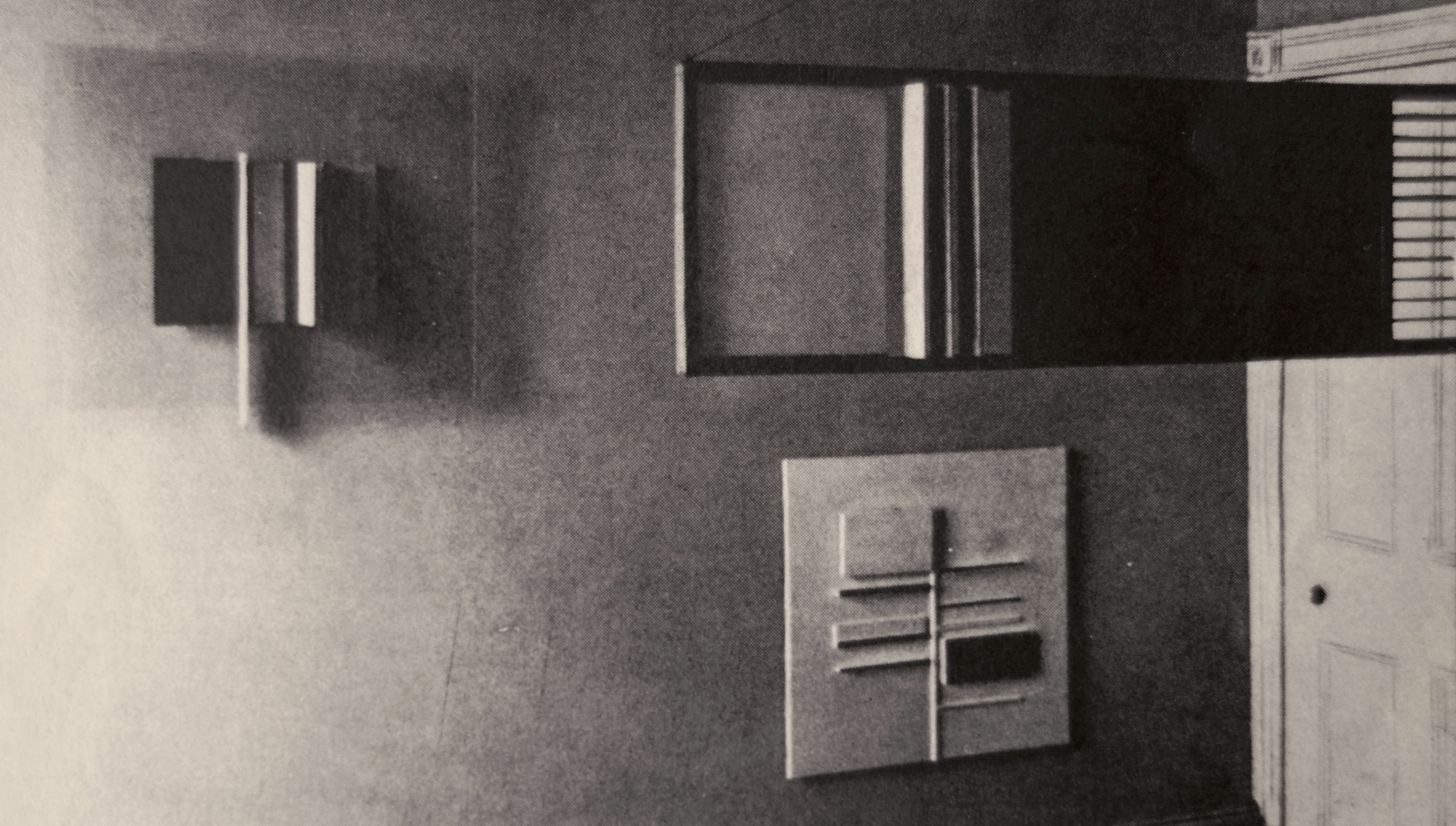 Installation photograph. Abstract works of art comprising stacked panels in rectilinear geometric designs are arranged on and in front of a wall in this black and white photograph.