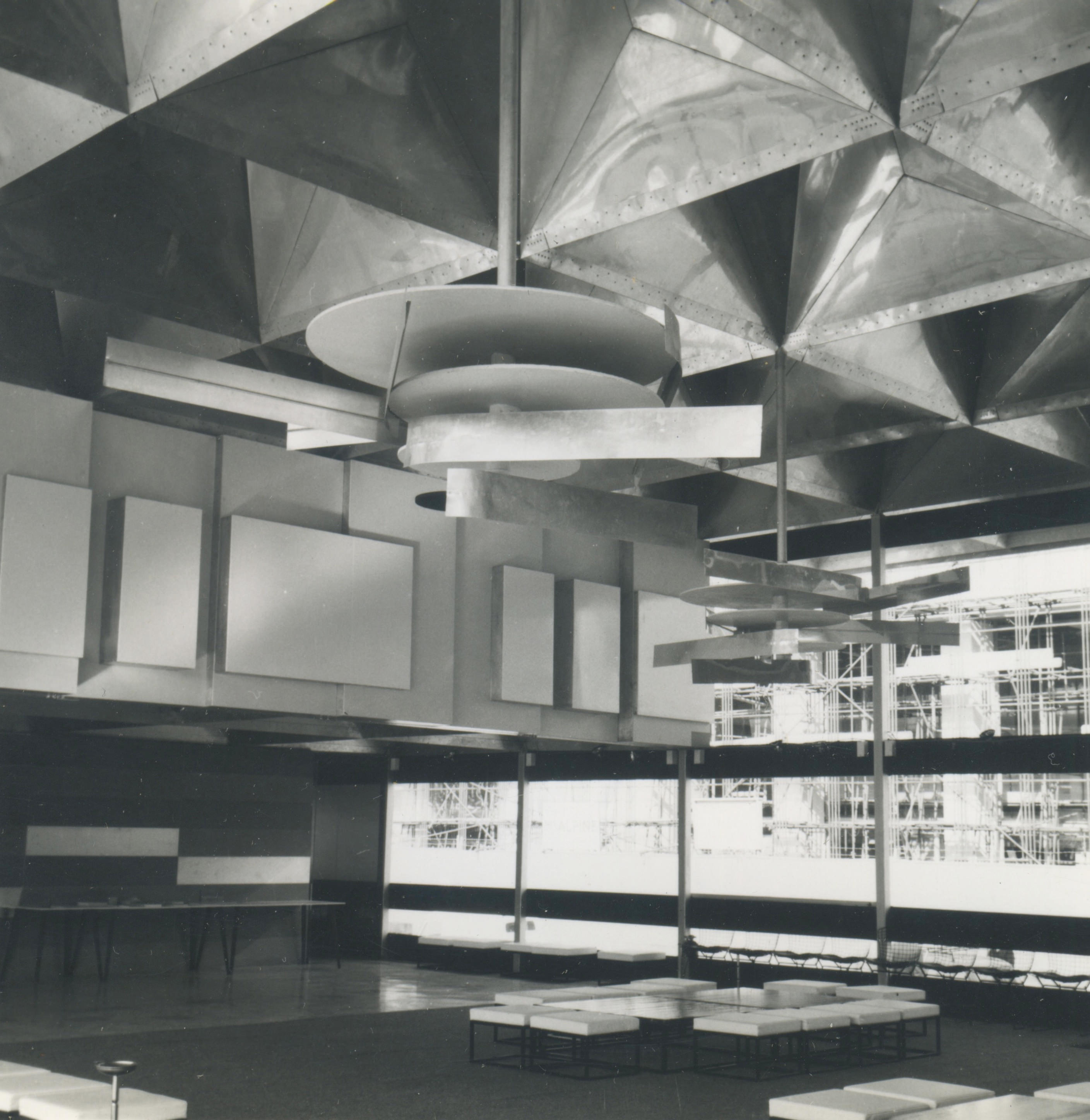 Installation photograph. Black and white photograph of a window-lined room with deep, angular coffers in the ceiling above sleek-lined furniture. Two abstract sculptures hanging from the coffers are made up of flat disks and long strips of metal.