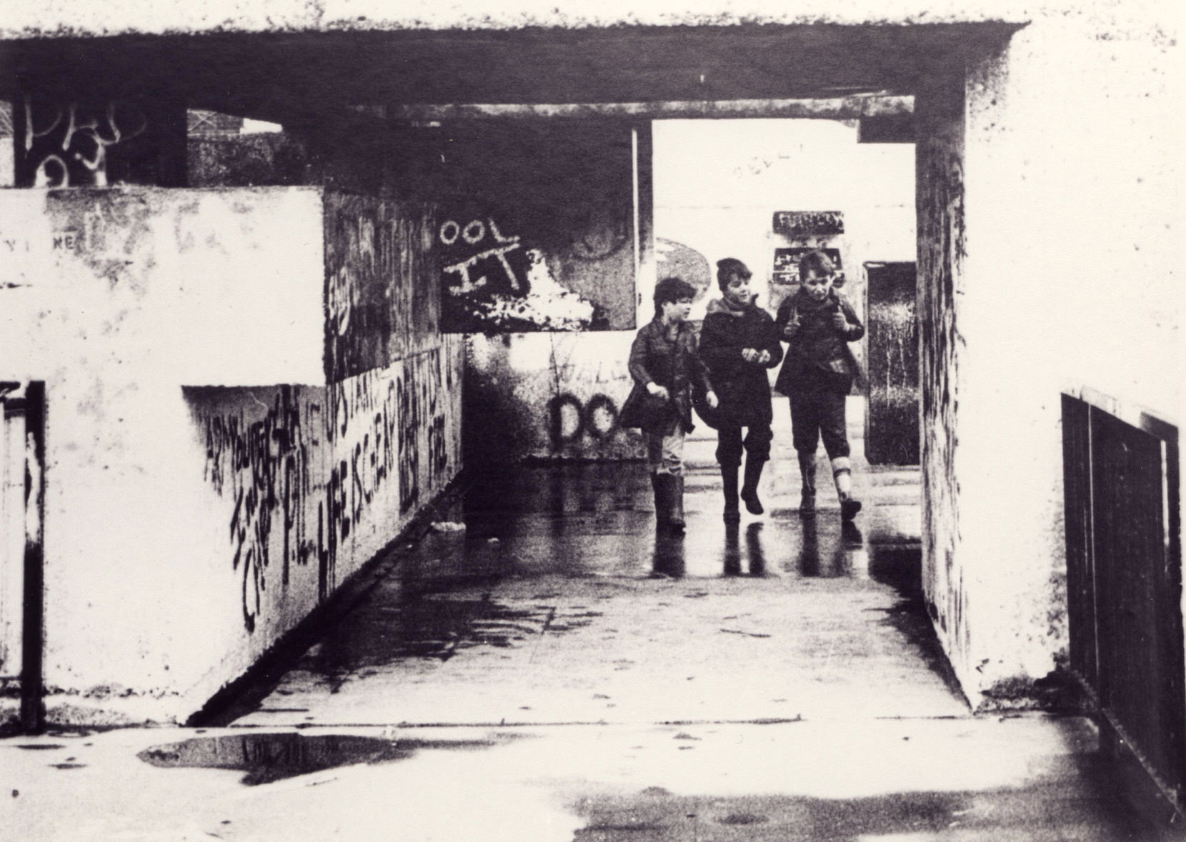 Architecture. Three boys walk towards us through a squared concrete opening lined with graffiti in this black and white photograph.
