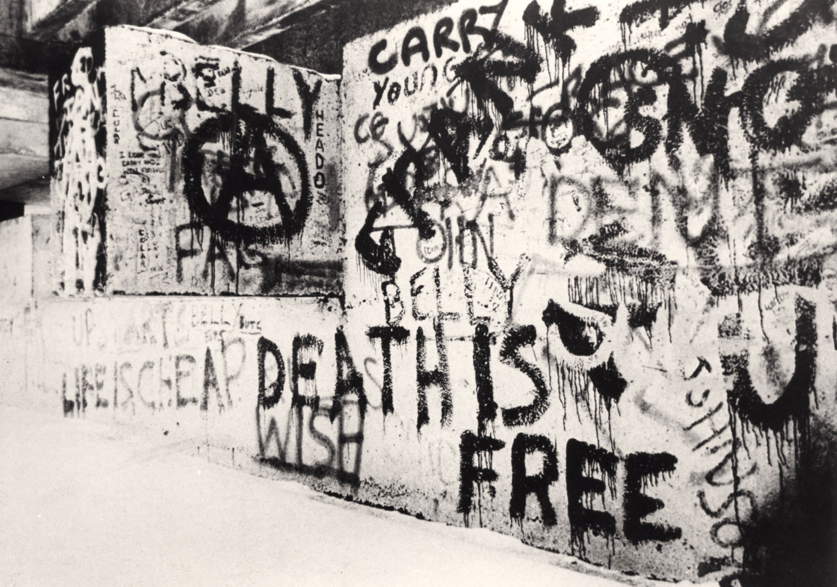 Architecture. A concrete wall is covered with graffiti in this black and white photograph. Some painted words and phrases read “Life is Cheap Death is Free”, “Wish”, “Belly,” and “Young”. A letter “A” within a circle is to our left.