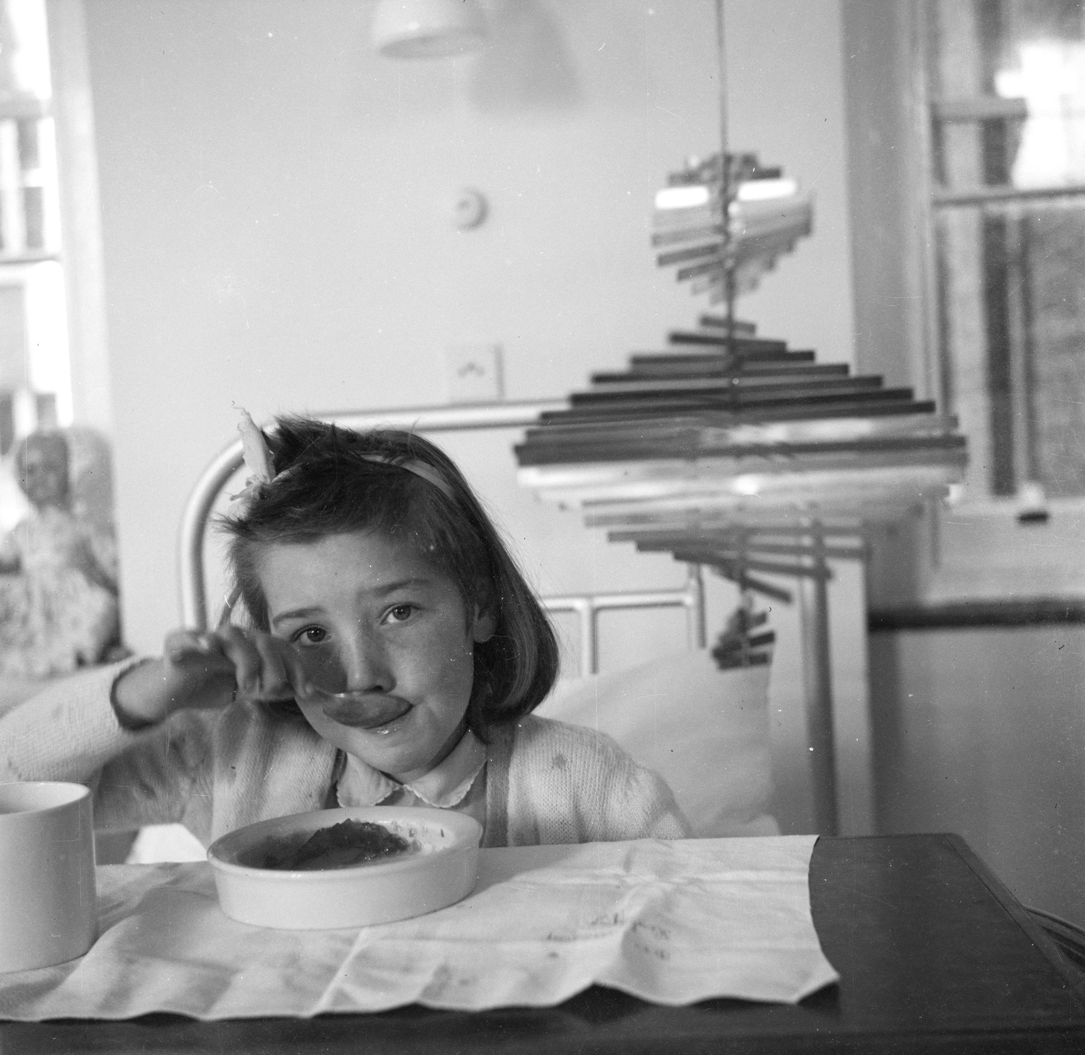 Black and white photograph. A child eats from a chest-high table close to us in this black and white photograph. The headboard of a hospital bed is visible behind her. A mobile of metal strips that increase and decrease in length to create a reverse hourglass shape hangs between the girl and the headboard.