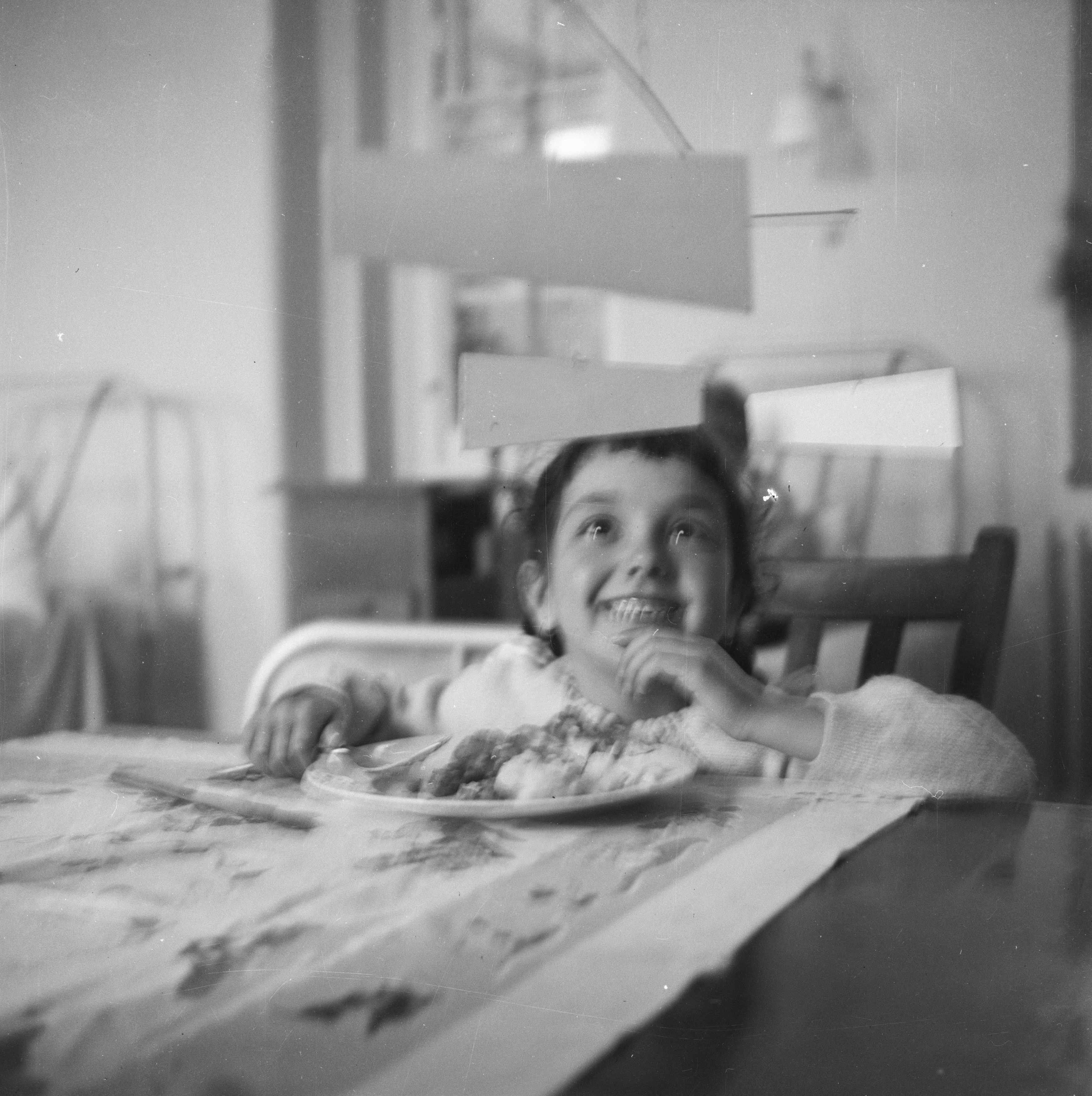 Black and white photograph. A child looks up at a mobile and grins broadly in this out-of-focus black and white photograph. A plate of food sits on a table at nearly chin-height on the child. Rectangular panels of the mobile float above.