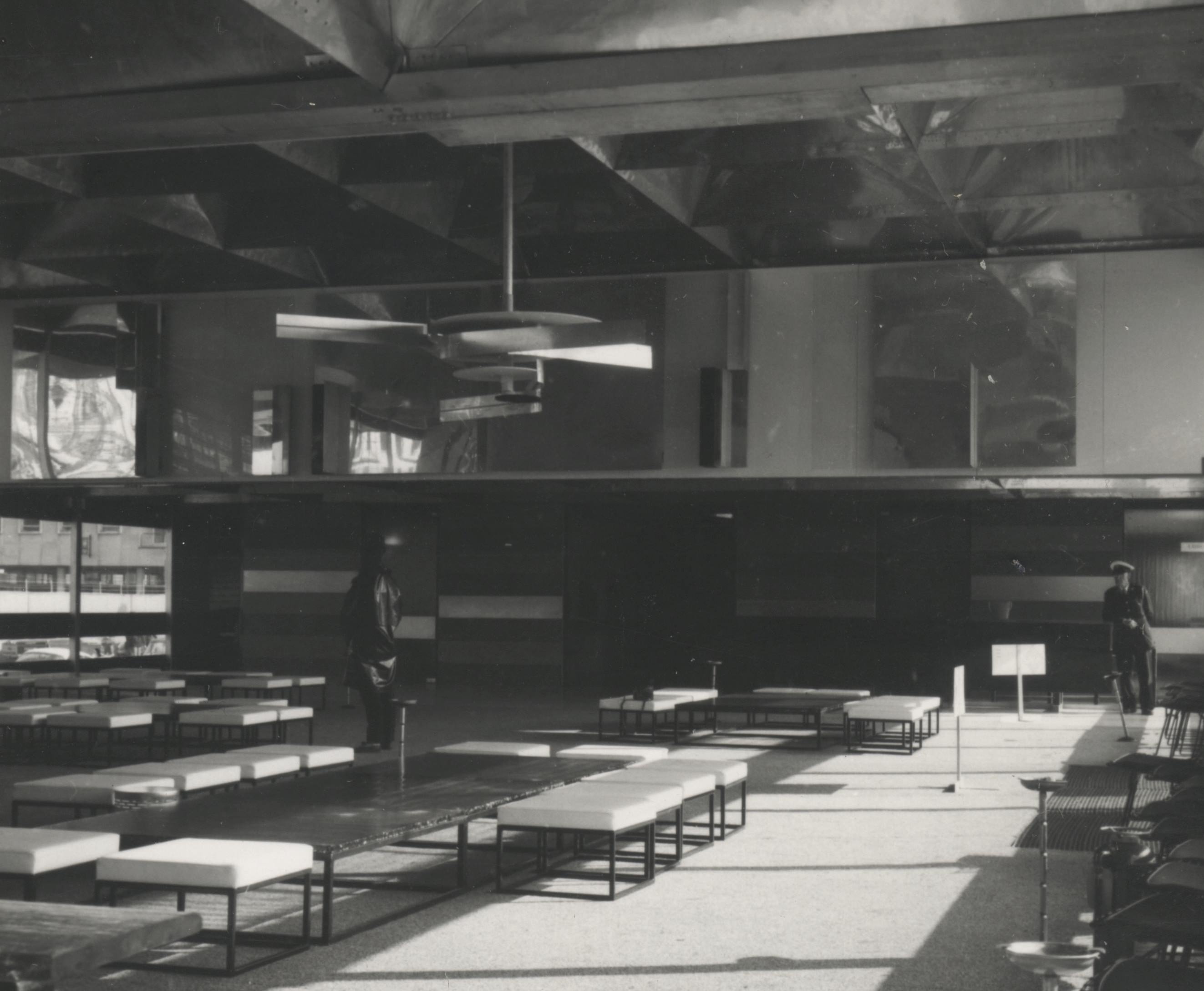 Installation photograph. Black and white photograph of a two-storey-high open space. An abstract sculpture made of discs and strips of metal hangs from the ceiling. On the far wall, the lower level is filled with horizontal bands in different shades. Reflective surfaces are above, on the second level of the open space.