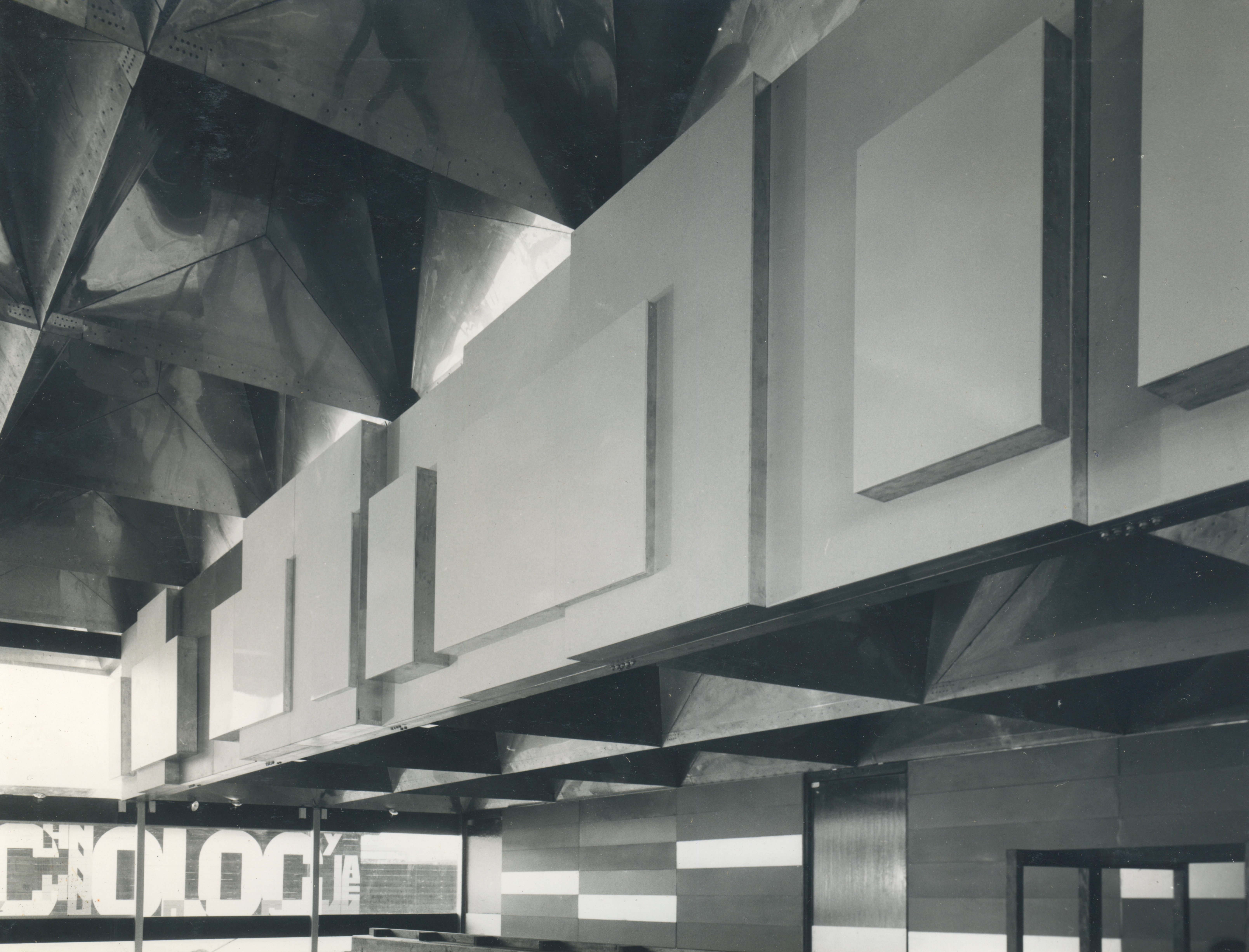 Installation. Black and white photograph of a two-storey-high open space in a building with a wall of windows opposite us. A frieze of rectangles of different sizes overlapping at different depths is along the upper storey.