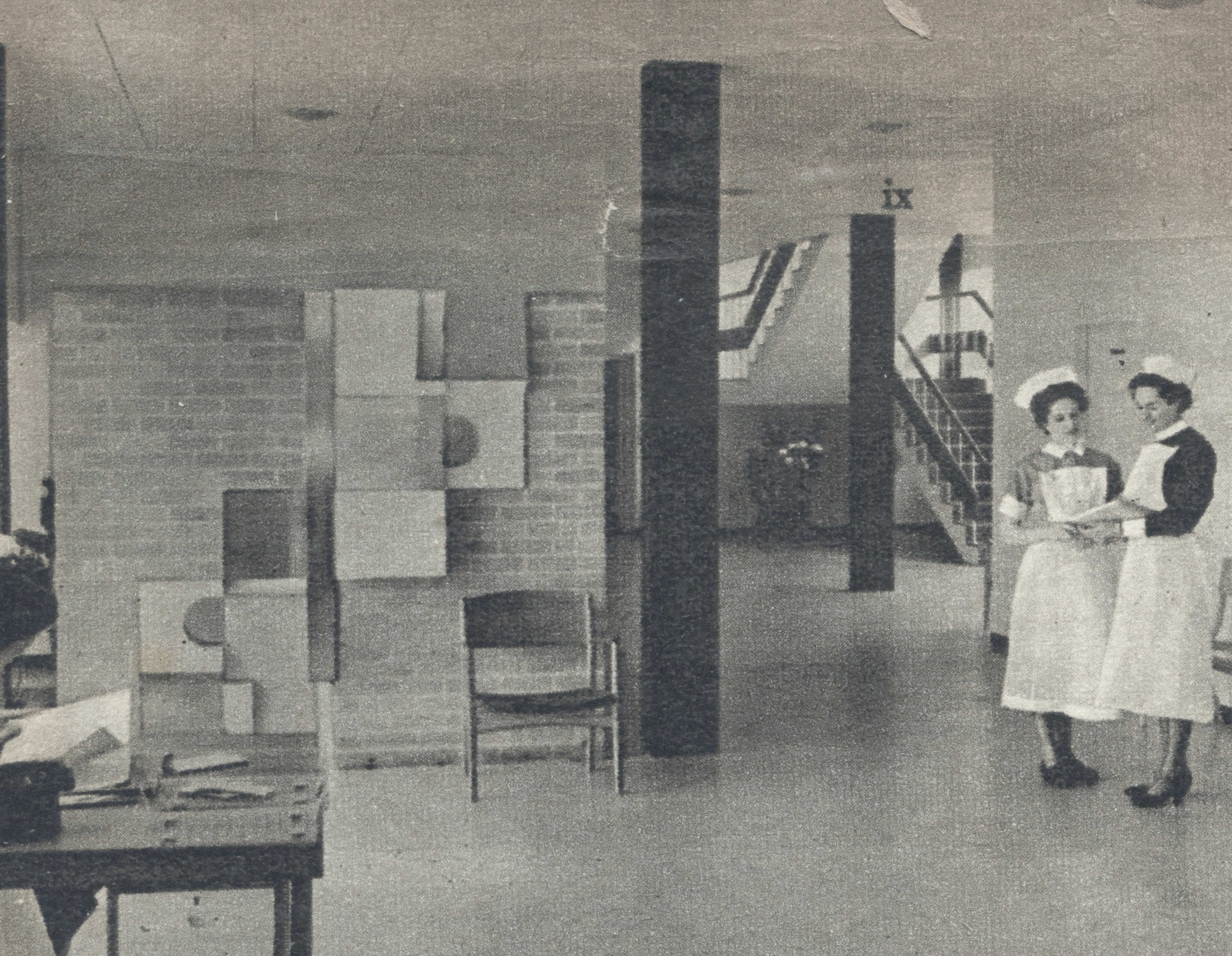Black and white photograph. Two uniformed nurses stand to one side of an open space with staircases at the back corner, to our right. To our left, square and rectangular panels create a cascading form against a brick wall.