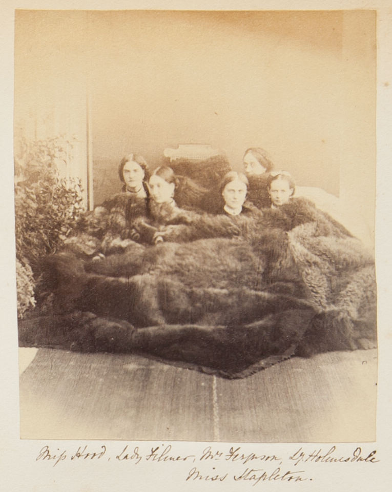 Black and white photograph. Five women with pale skin sit under a fur pelt so large it covers them all, so only their heads are visible. The women are identified in handwriting below.