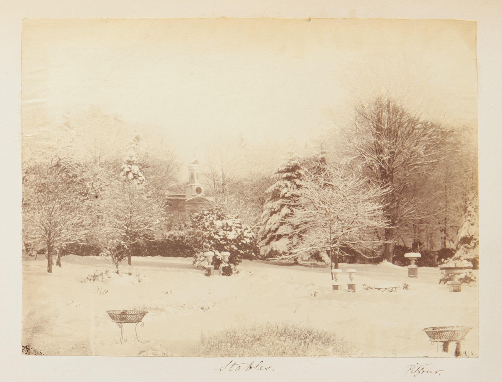 Black and white photograph. A snow-covered lawn leads back to trees lining a lane. A steeple-like tower is nestled in the trees along the horizon. Handwriting below reads “Stables” and “Pitfour”.