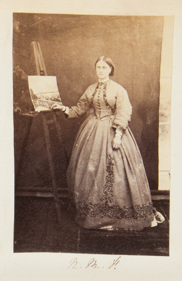 Black and white photograph. A pale-skinned woman wearing a long-sleeved dress with a full, wide skirt stands with one hand resting on the edge of an easel, which holds a painting.