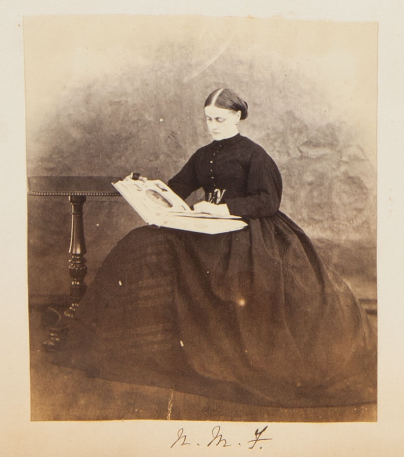 Black and white photograph. A pale-skinned woman wearing a long, puffy-sleeved dress with a full, wide skirt sits looking at an album. Her dark hair is parted down the middle and pulled back at the nape of her neck. A side table sits next to her right hand, to our left.