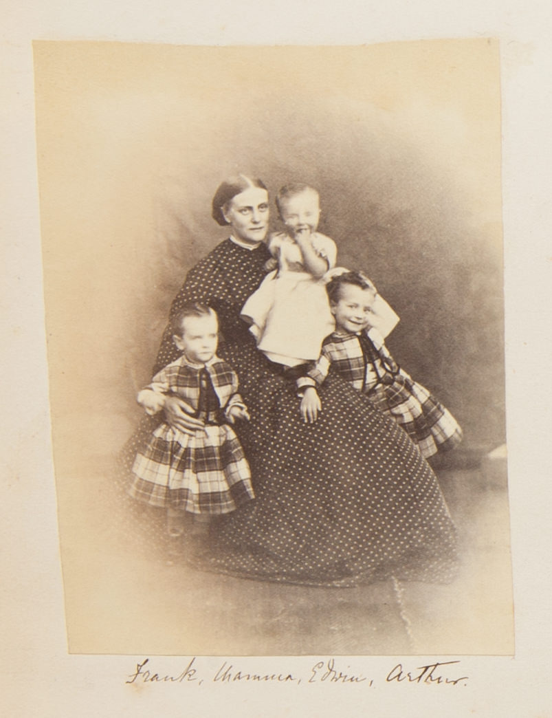 Black and white photograph. A pale-skinned woman wearing a dark dress with white polka dots sits and holds a baby as two small children lean against her lap. The standing children wear plaid garments and one of the smiles at us. The baby holds one fist in their mouth and also smiles.