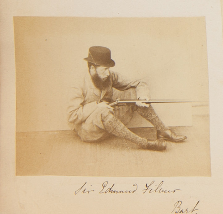 Black and white photograph. A man with pale skin and a full, bushy beard sits on the ground, knees bent so he can steady a rifle. He faces our right almost in profile. His name is written in pen below.
