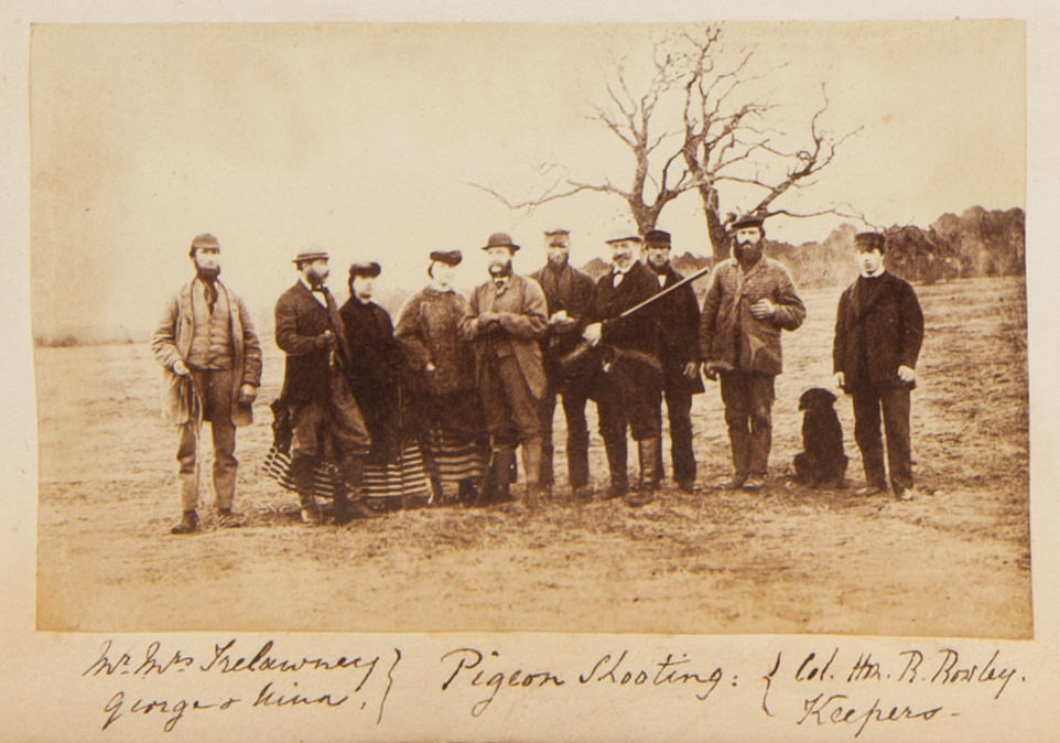 Black and white photograph. Two women, eight men, and a dog stand in a row, posing in front of a flat landscape. One man holds a long rifle. The kinky branches of a leafless tree just behind the group twist against a blank sky. The people are identified in pen along the bottom of the image to either side of the title, “Pigeon Shooting”.