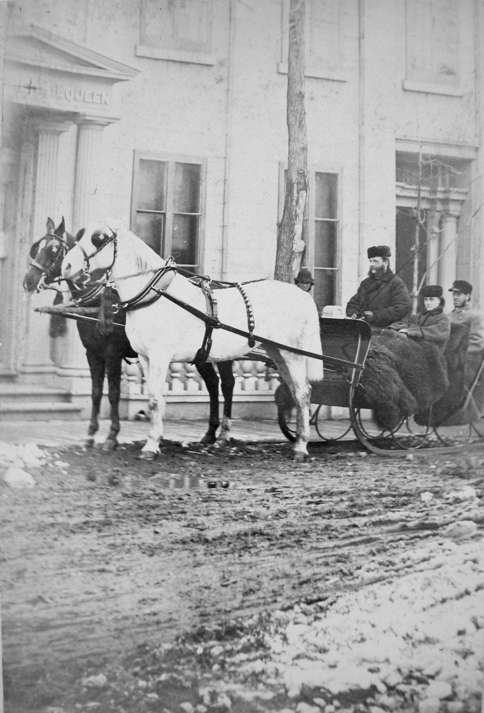 Black and white photograph. Two people with furs draped over their laps sit in the back of a horse-drawn sleigh. The driver in front holds a whip, and another person looks towards us from behind the horses. All the people have pale skin and wear hats and coats.