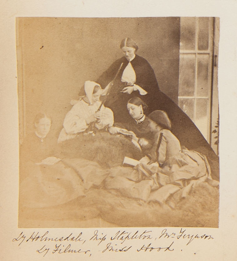 Black and white photograph. An enormous fur blanket is draped over one woman’s lap and across the ground. Three more women sit on the fur, reading books. The woman with the fur over her legs holds a long-barrelled gun angled upwards. A woman in black is at the apex. The women are identified in handwriting below.