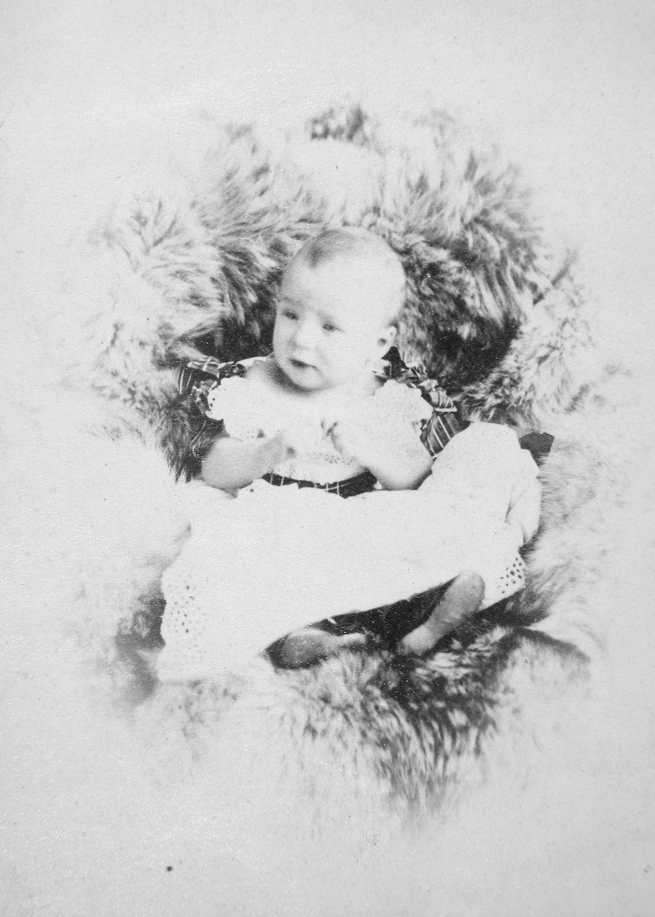 Black and white photograph. A pale-skinned baby wearing a white dress-like garment and shoes lies cushioned in a fur pelt. The baby’s features are blurry, but he looks off to the left, head and hands lifted.