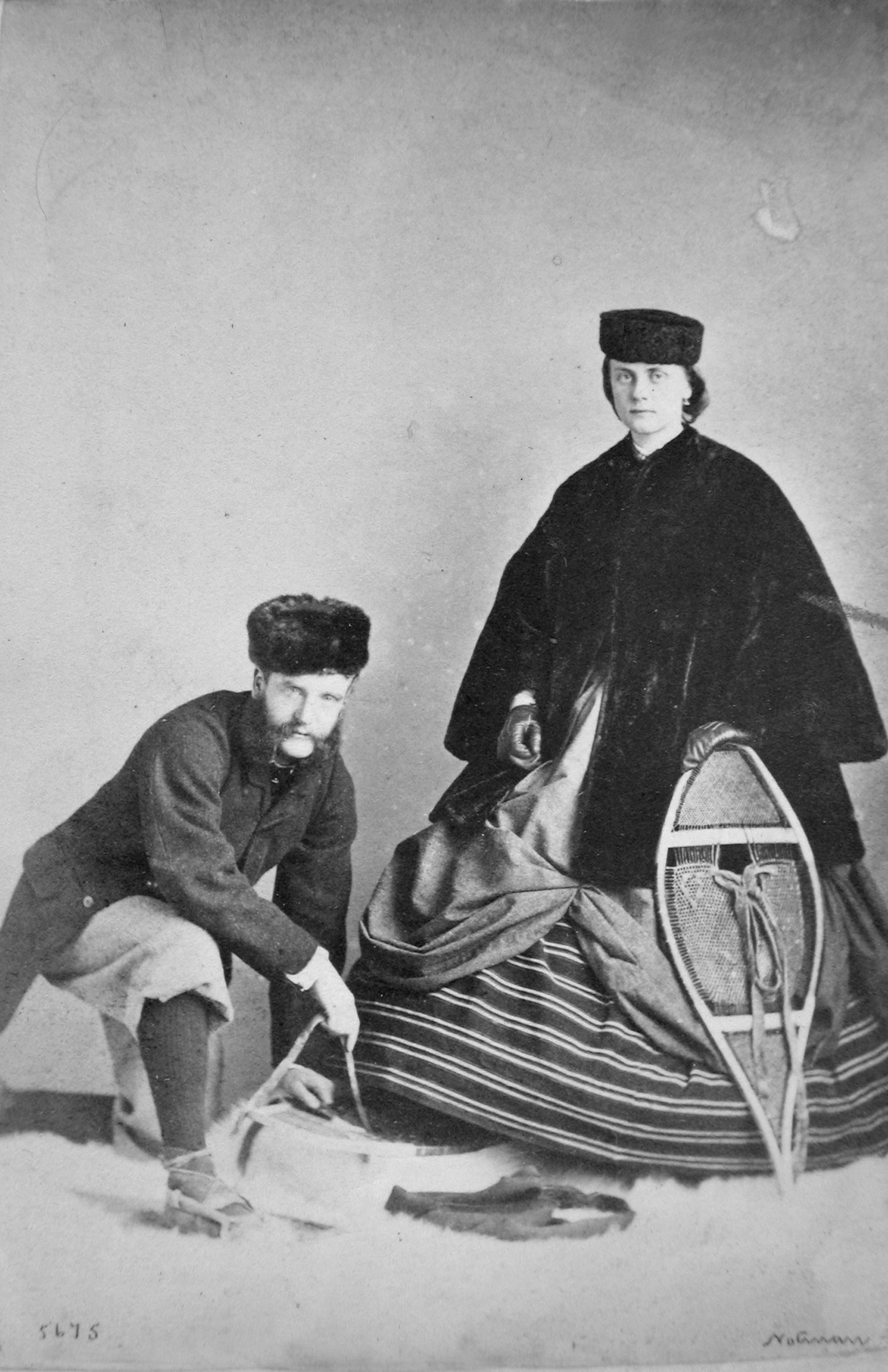 Black and white photograph. A man and woman pose in a studio. They both have pale skin and wear fur caps and heavy coats. The woman stands with a snowshoe propped against her hoop skirt. The man kneels, holding the rope for what might be a sled.