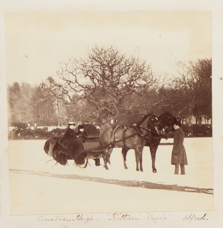 Black and white photograph. Two dark horses stand ready to pull a sleigh occupied by two women. A man wearing a long coat stands at the horses’ heads. Leafless trees are lacy against a blank sky over an expanse of white, snowy lawn. The title and peoples’ names are written in pen below.