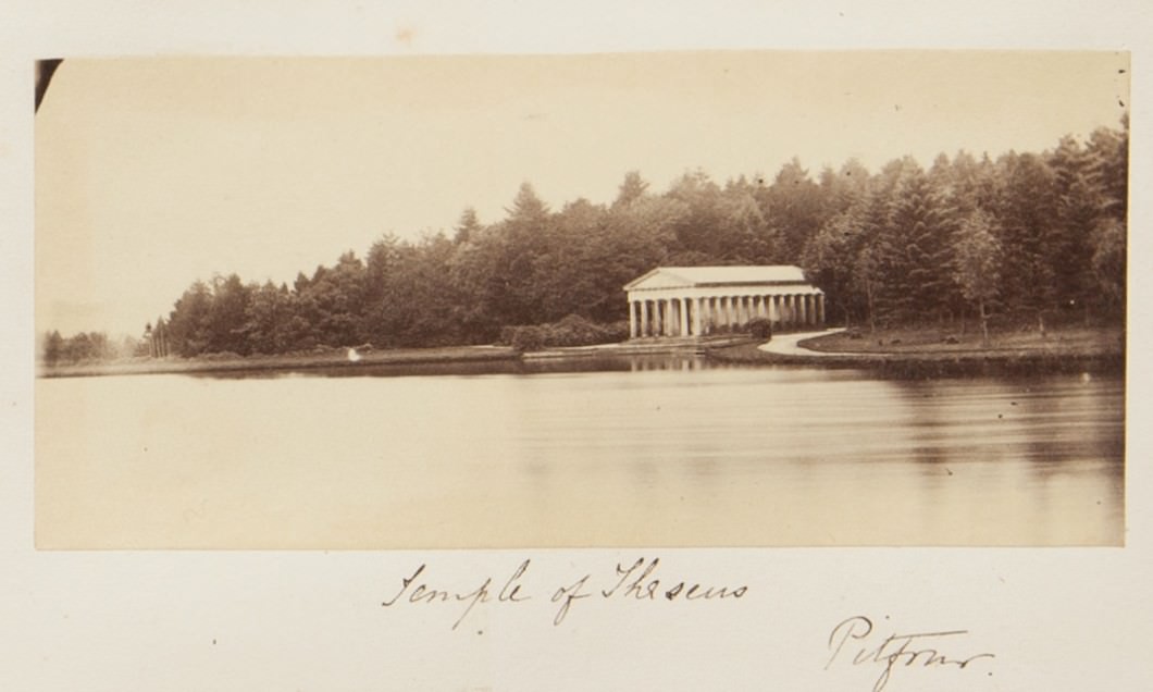 Black and white photograph. We look across a lake at a rectangular, columned building with a peaked roof nestled among evergreens. The surface of the water appears smooth and bright, like the sky above. The title is written in pen below, along with “Pitfour”.