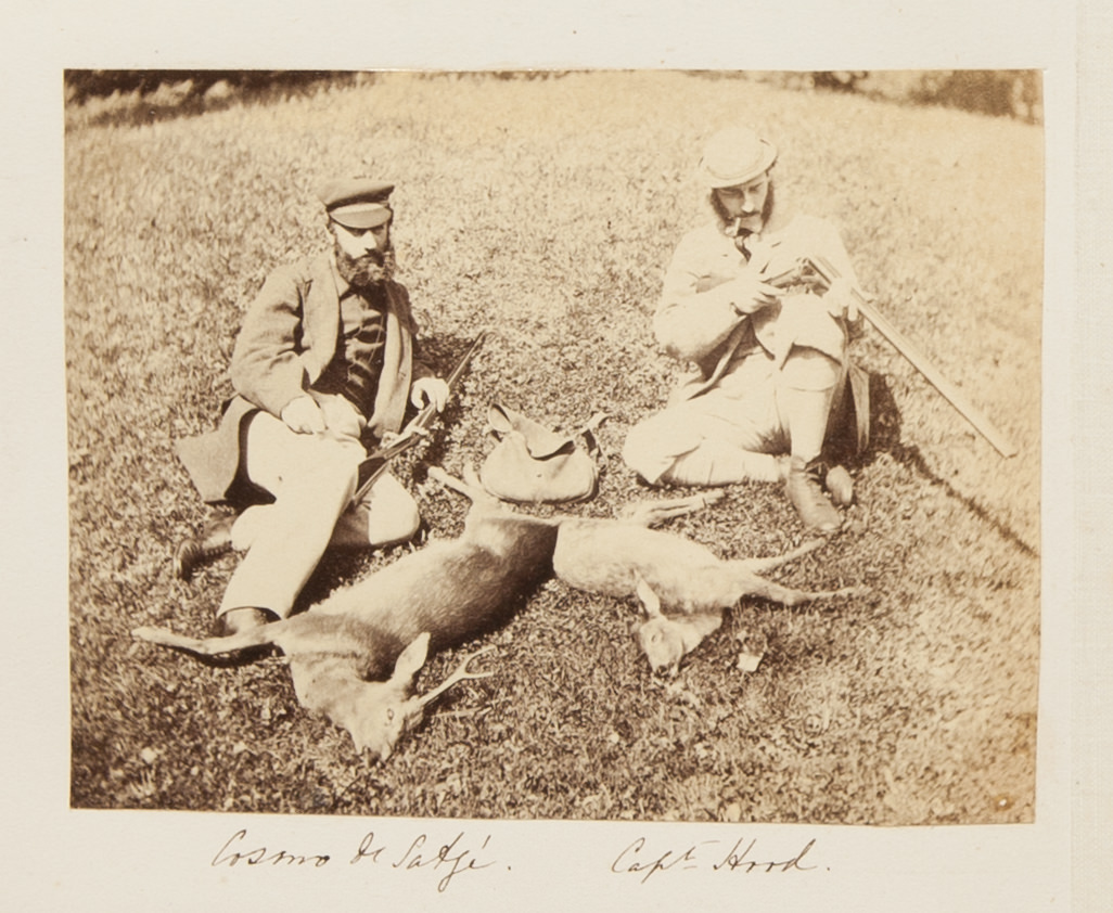 Black and white photograph. Two pale-skinned men recline in the grass above two dead deer. The men have rifles and wear caps and suits with vests. The man to our right has a clay pipe in one corner of his mouth. The men’s names are written in pen below.