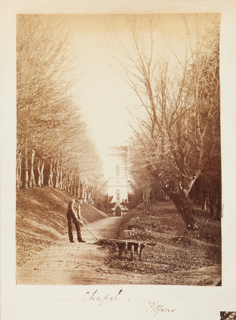Black and white photograph. A man clears fallen leaves from a lane. Walls of trees grow to either side of the path, which leads back to a crenelated tower. The location is handwritten in pen below, “Chapel” and “Pitfour”.