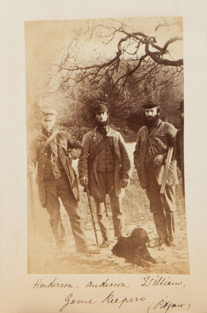 Black and white photograph. Three men stand under a leafless tree in this vertical composition. They wear caps and coats criss-crossed with straps. A dark dog lies at their feet. Their names are written in pen below: “Henderson, Andersson, William, Game Keepers” and, in parentheses, “Pitfour”.