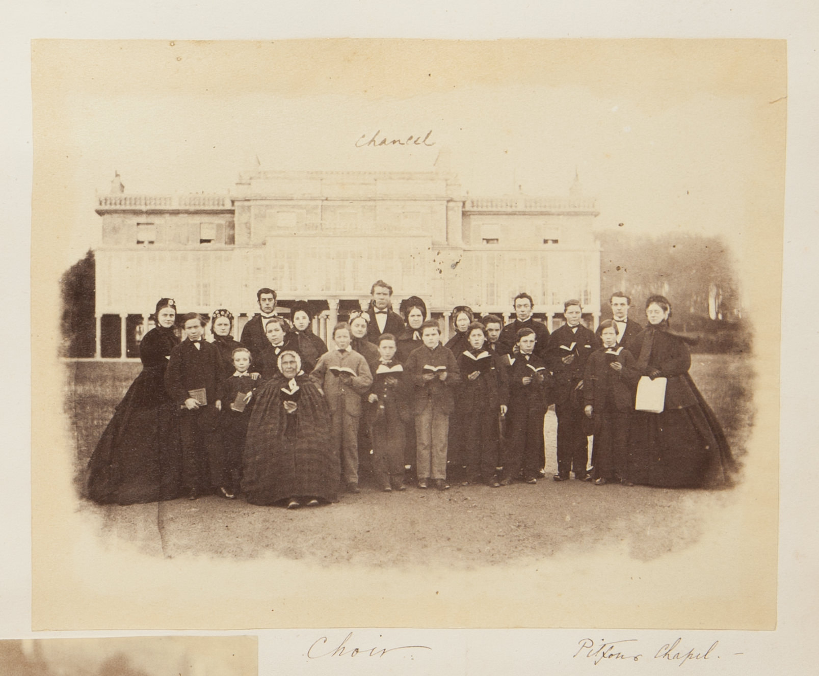 Black and white photograph. Twenty-three men, women, and children stand or sit, most holding choir books the size of paperback novels open, in front of a mansion. They all have pale skin. The title is written in pen below along with the location, “Pitfour Chapel”.