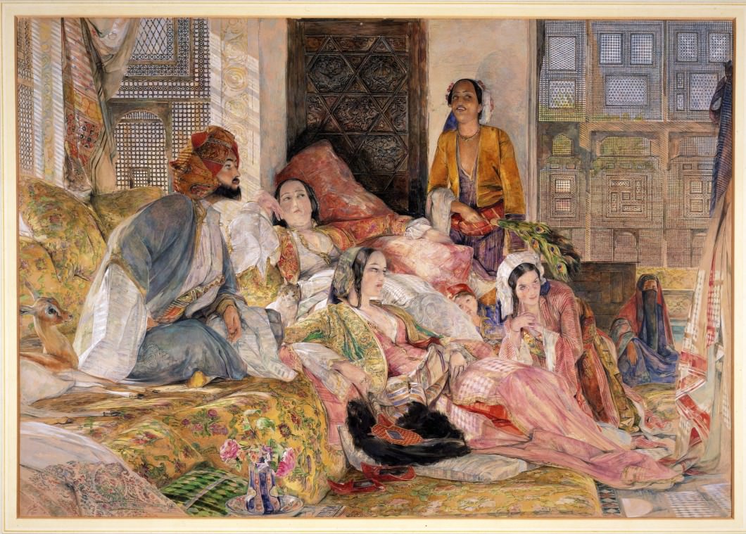 Watercolour. Four women, two men, and a child look at something out of our view to the right. One man sitting cross-legged on a couch wears an elaborate turban and voluminous robes. Three of the women recline and lounge near him. The women have pale skin and the two men more tanned complexions.