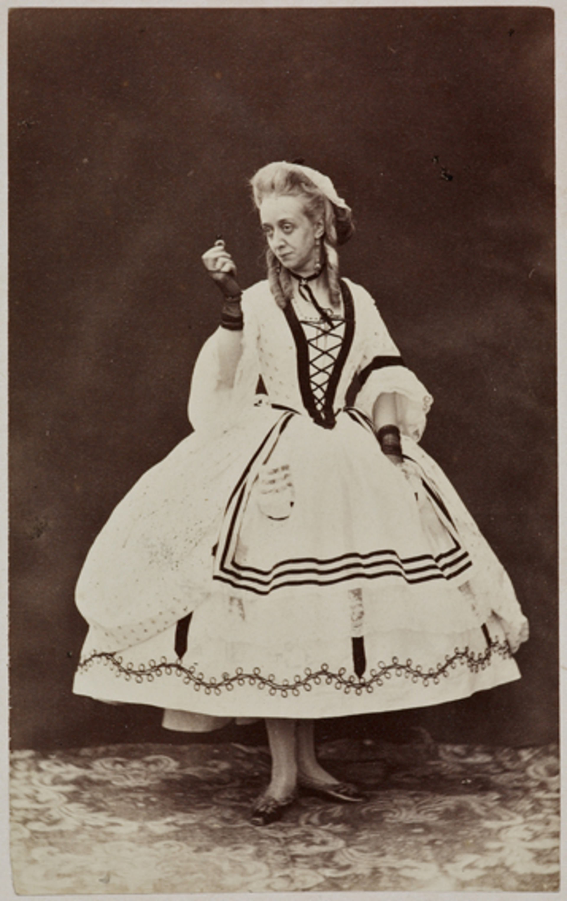 Black and white photograph. A pale-skinned woman wears a white dress edged with black lines or designs. The skirt flares out from the hips and ends above the ankles. She holds up a ring with her right hand, to our left, and looks in that direction against a dark background.
