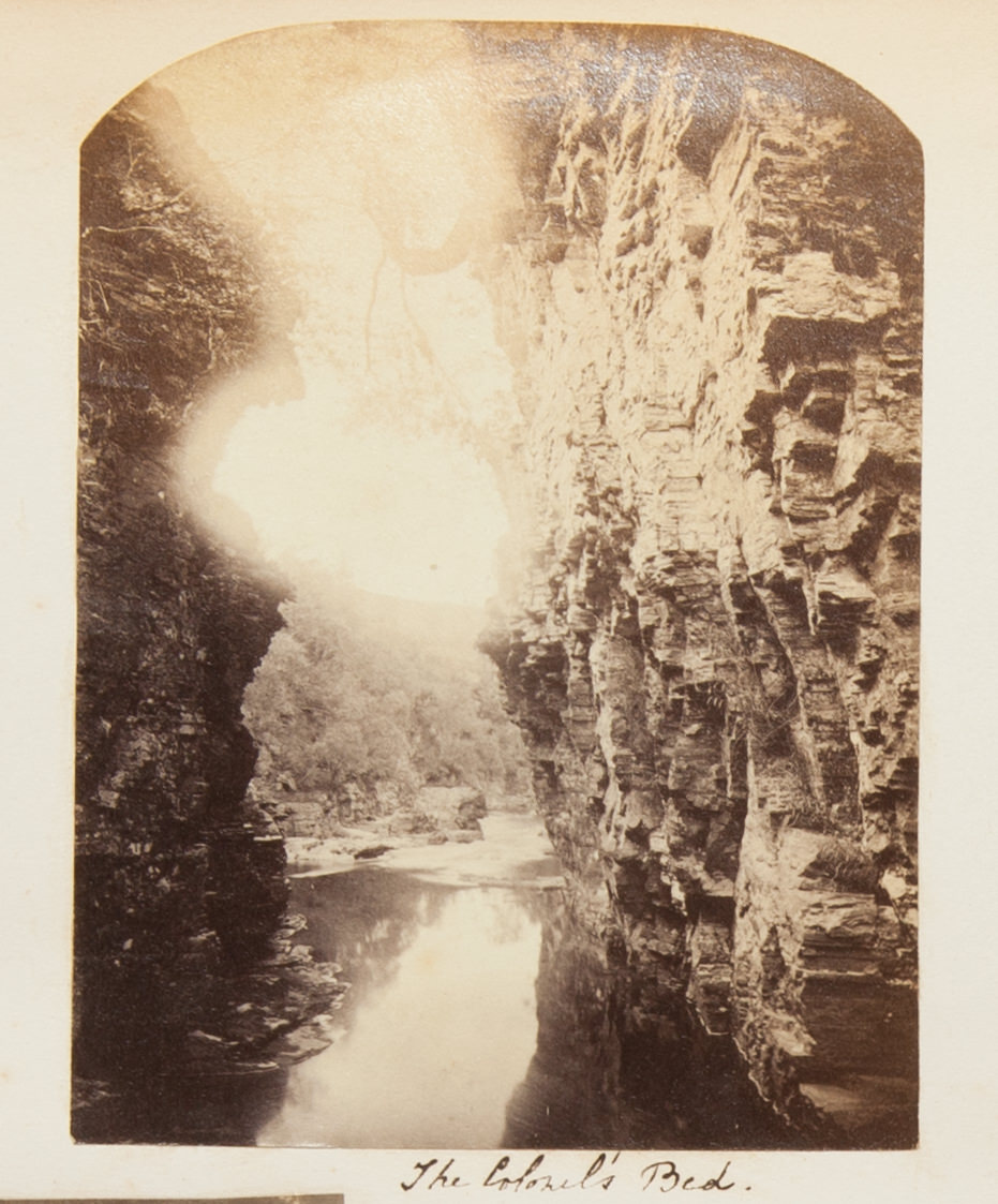 Black and white photograph. Sheer, rocky cliffs tower up both sides of this photograph. The river running between them and away from us appears to have a smooth surface. Sunlight washes out the top centre of the photograph, over a tree-lined hill. The title is written in pen below.
