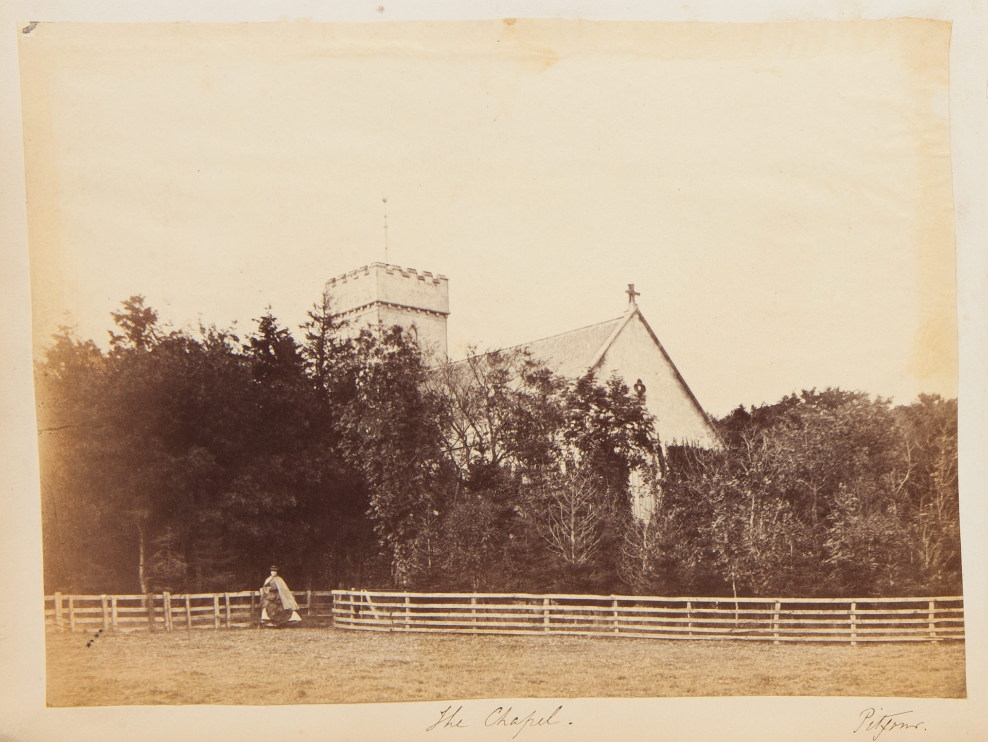 Black and white photograph. A woman wearing a hooped skirt under a cape stands at a fence that lines a grassy field. Beyond the fence is a line of trees, above which the crenelated tower and peaked roof of a church are outlined against a blank sky. The title is written in pen below and, to our right, “Pitfour”.