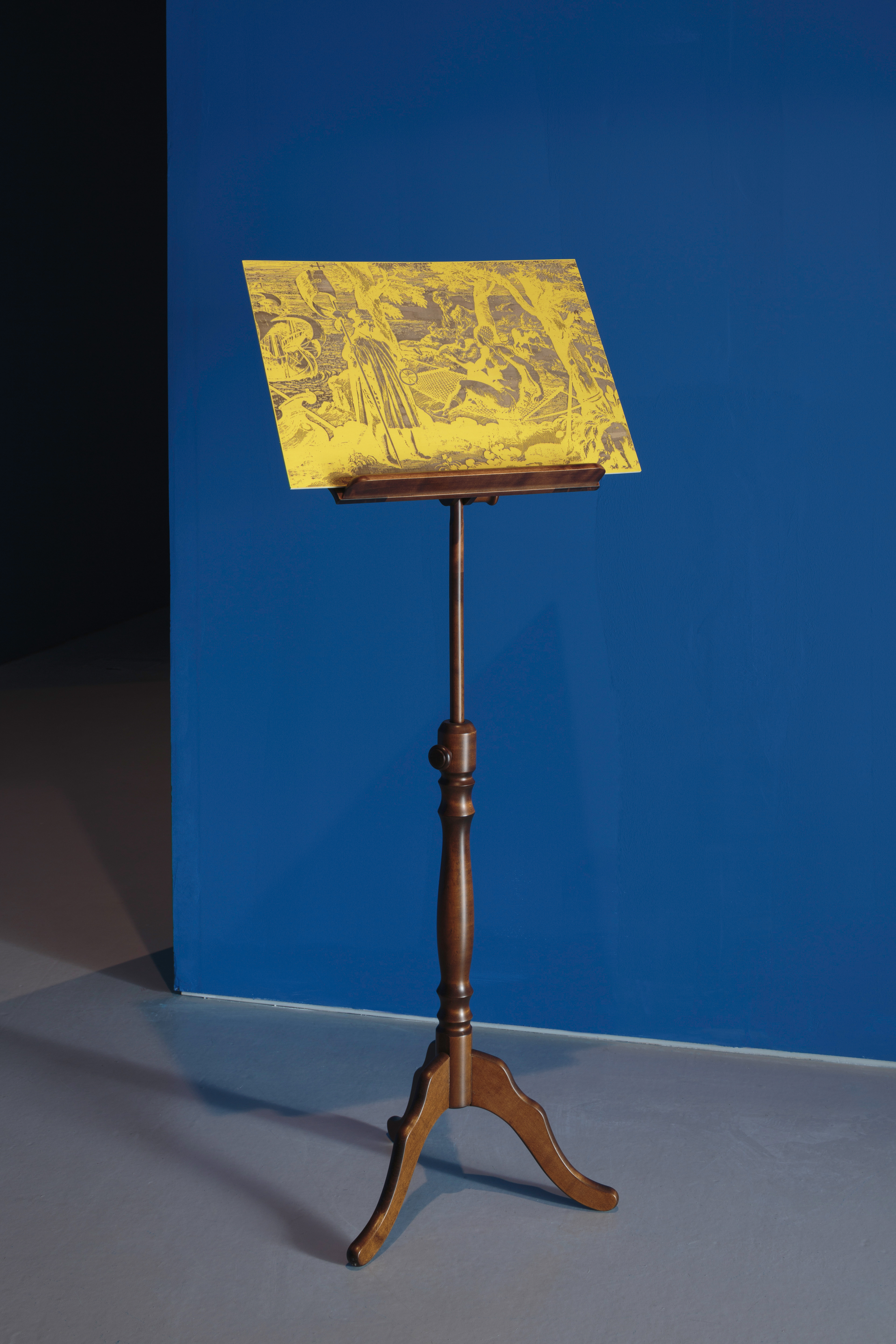 Installation. A gold-coloured scene is displayed on a wooden music stand against a gallery wall painted cerulean blue. The gold scene resembles a photographic negative. Though details are difficult to make out, a man wearing a calf-length robe stands in front of what appears to be a nude woman reclining in a forest. A ship on rippling waters is to our left.