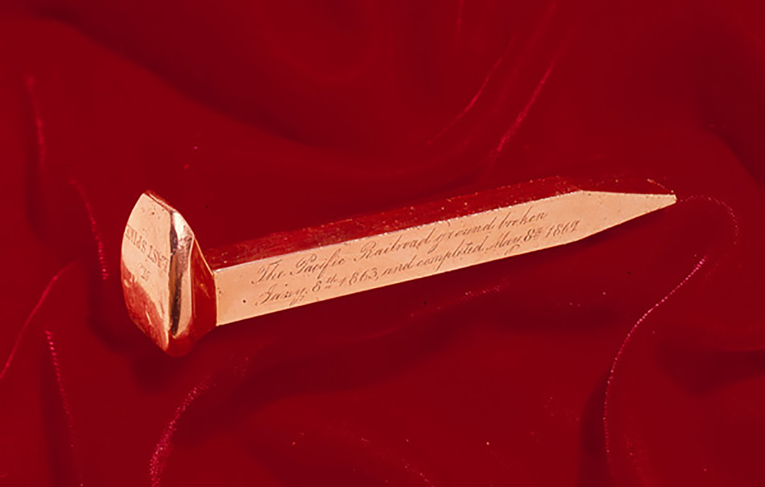 Golden railroad spike is engraved on the long side we can see as follows: “The Pacific Railroad ground broken January 8th 1863, and completed May 8th 1869”. It lies on scarlet-red fabric, perhaps velvet.