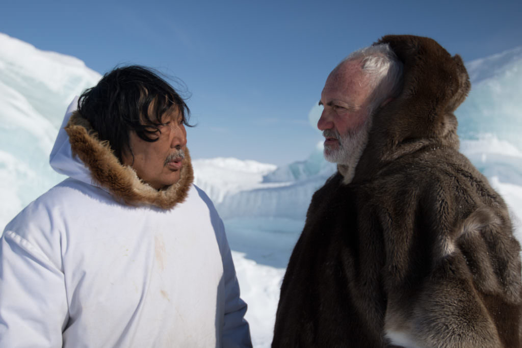 Video still. Two men stand near us. To our left, a man wears a white parka with a fur-lined hood. He has dark skin, black hair, and a grey moustache. He talks to a pale-skinned man, to our right. The latter has a white beard and hair, and wears a fur parka with the hood up. They stand in front of a snowy expanse.