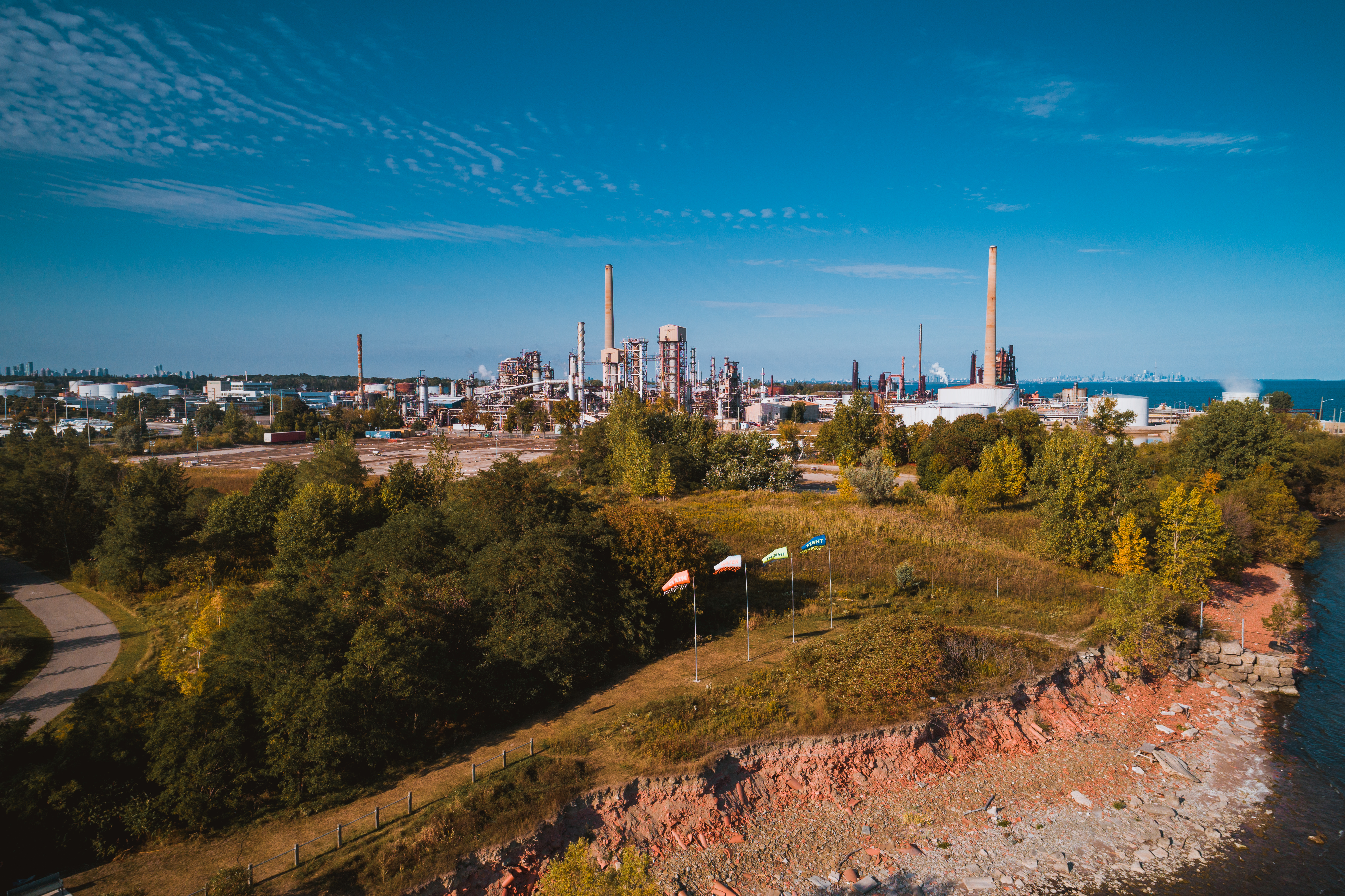 Public art. From an aerial perspective, we look down onto four windsocks planted on tall flag poles near a rocky shoreline. An industrial area is beyond, with smokestacks outlined against the vivid blue sky.