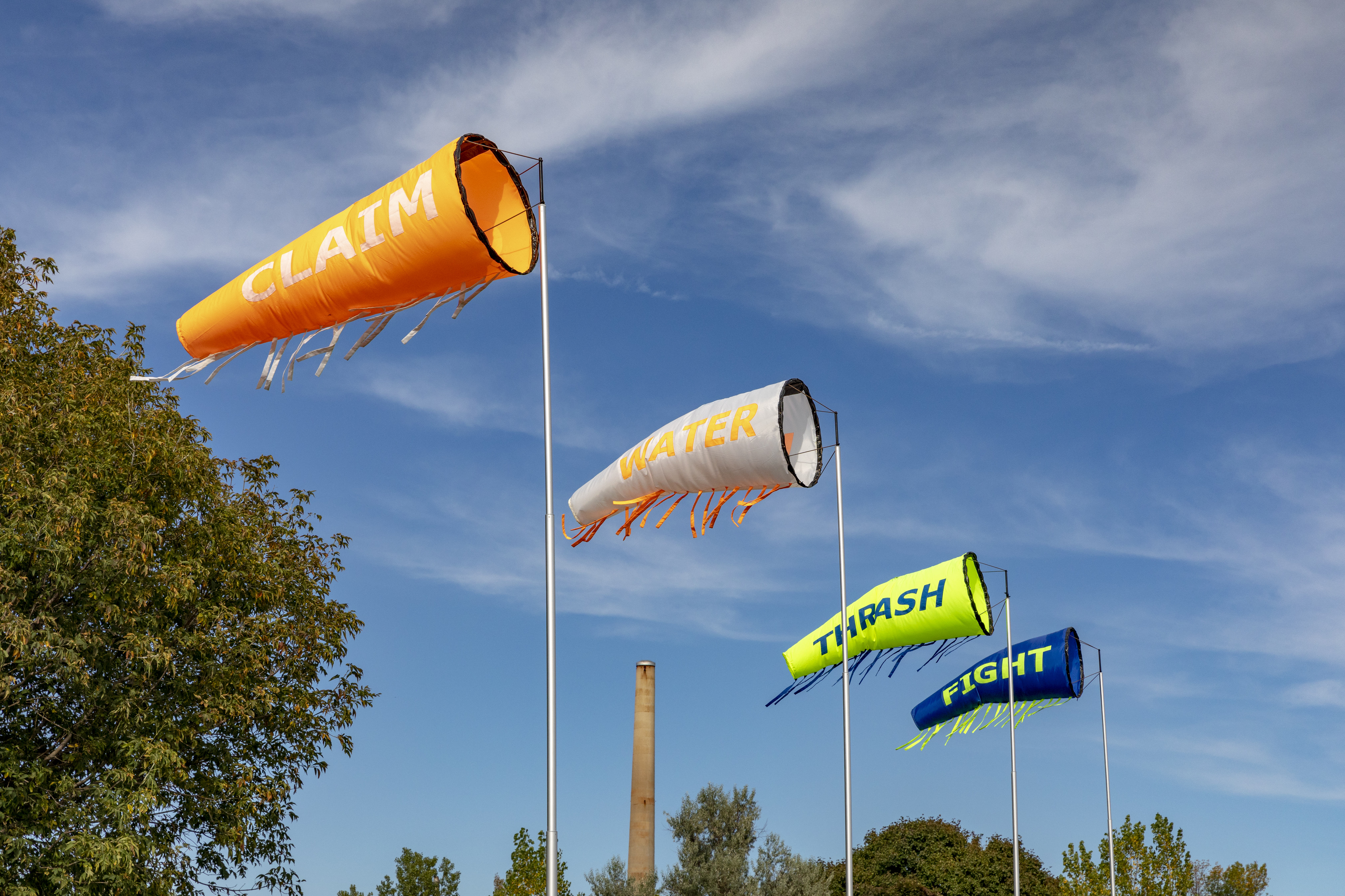 Public art. Four windsocks fill with air so a single word on each can be read. In white letters against an orange sock, the one closest to us reads “Claim”. In orange against white, the next reads “Water”. Then in blue against neon green is “Thrash” and, finally, in green against blue, “Fight”.